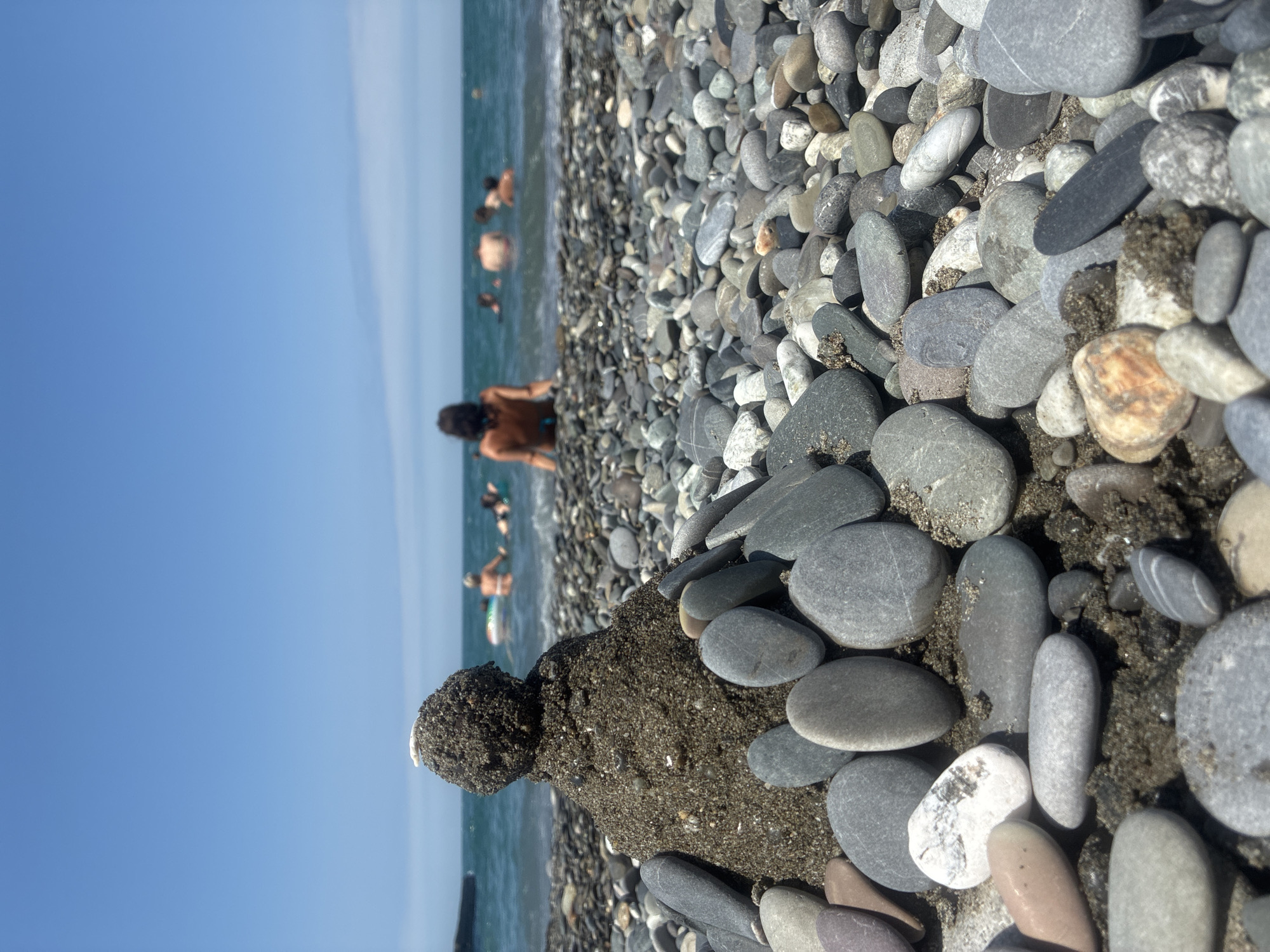 A low-angle, close-up shot of a small sandcastle adorned with stones on a pebbled beach in Sochi, Russia. The sandcastle is situated in the foreground, with its base surrounded by various grey and white pebbles. The ocean is visible in the background, with several people swimming and relaxing in the water under a clear blue sky. The overall atmosphere is one of a sunny day at the beach.