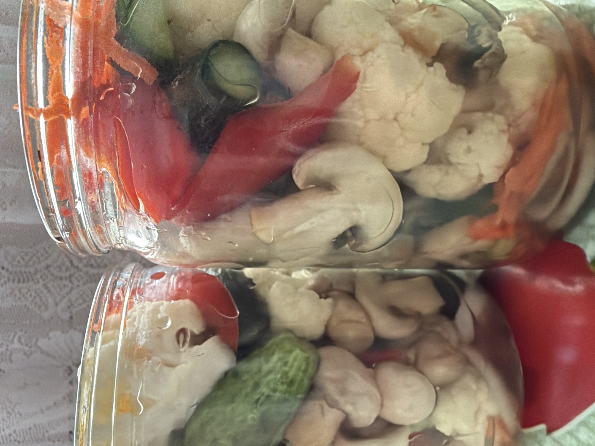 Two glass jars filled with pickled vegetables are shown. The jar on the left contains cauliflower, cucumbers, and what appears to be white beans or mushrooms. The jar on the right contains cauliflower, red peppers, cucumbers, carrots, and mushrooms. The jars are packed tightly with the pickled ingredients submerged in liquid. The background is a blurred light-colored lace-like fabric. The image is a close-up shot, with a shallow depth of field. The setting is indoors, and the lighting appears to be natural. There are no people present in the image.