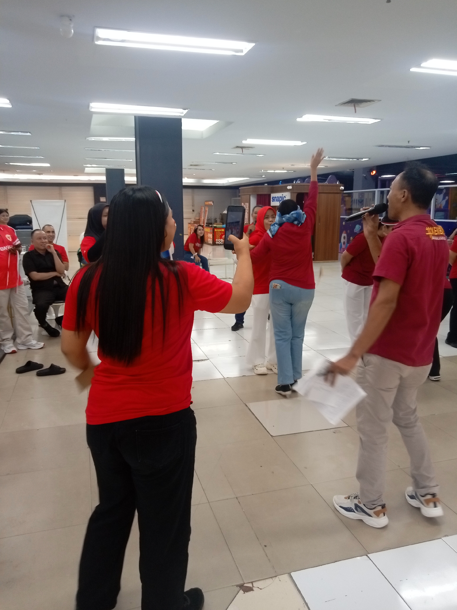 The image captures an indoor gathering, possibly at a community center or large retail space, within the city of Batam, Indonesia. The scene is brightly lit by fluorescent ceiling lights. Several individuals are present, most wearing red shirts. One woman in the foreground, facing away from the camera, has long black hair and is wearing a red t-shirt and black pants. She appears to be holding a smartphone and possibly filming or taking photos.
In the background, a man in a red shirt is holding a microphone and speaking, suggesting he might be an event organizer or performer. To his left, a woman in a red shirt and a blue headscarf is gesturing with her arm raised. Several other people in red shirts are visible, some seated and others standing. The floor is tiled with a beige and white checkered pattern. In the background, there are structures that resemble shelving or display units, one with a sign that says "snapic". The overall atmosphere appears to be lively and social.