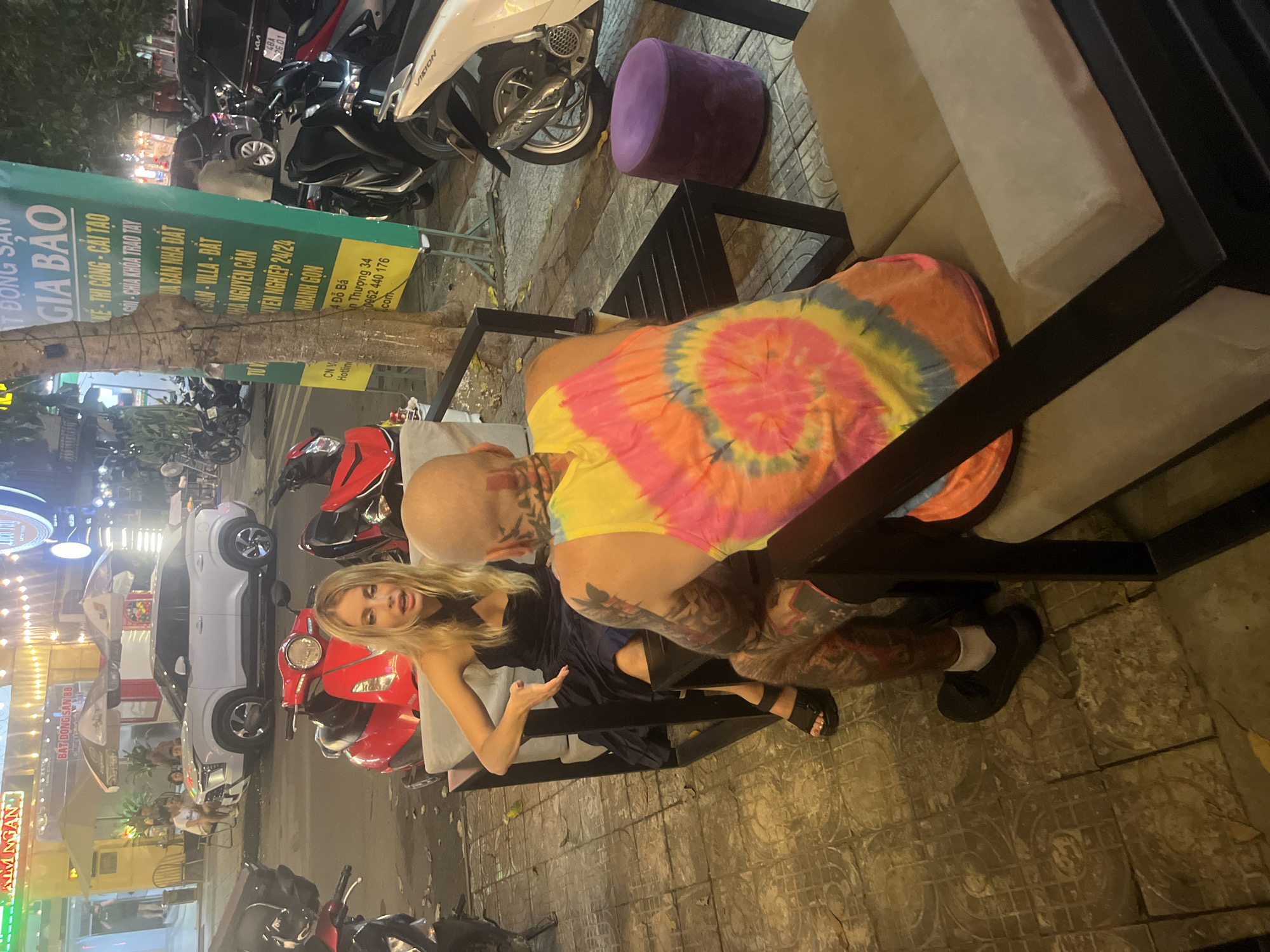 The image depicts an outdoor seating area at night, likely a cafe or restaurant, on a street in Đà Nẵng, Vietnam. A white SUV and several motorcycles are parked nearby on the street, suggesting an urban setting. A woman with blonde hair, wearing a dark dress, is seated and gesturing with her hands. She is sitting in a modern outdoor sofa, facing a man who is also seated and facing her. The man has a bald head and numerous tattoos covering his arms and visible on his neck. He is wearing a brightly colored tie-dye shirt. The seating area has a metallic frame with upholstered cushions. In the background, a large green sign with Vietnamese text is visible, indicating a business or advertisement. The street is paved with tiles, and fallen leaves are scattered on the ground. The overall atmosphere is casual and appears to be a social gathering or interaction taking place in the evening.