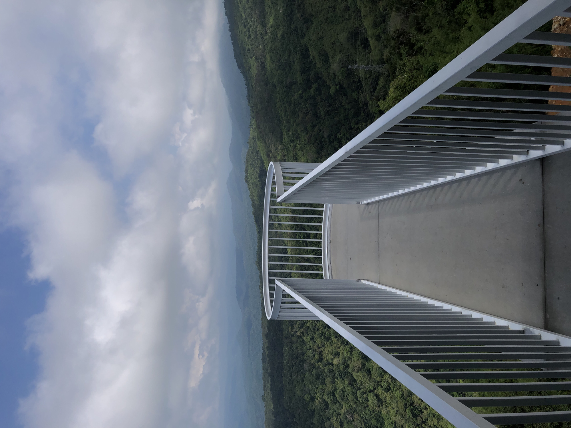 The image depicts a scenic overlook with a curved, white metal railing bordering a concrete pathway. Beyond the railing, a vast expanse of lush green hills and mountains stretches into the distance, partially obscured by a cloudy sky. The foreground is dominated by the overlook structure, creating a sense of depth and perspective. The scene suggests a natural landscape, likely a tourist attraction or a viewpoint. The location is specified as Nam Hải, Vietnam.