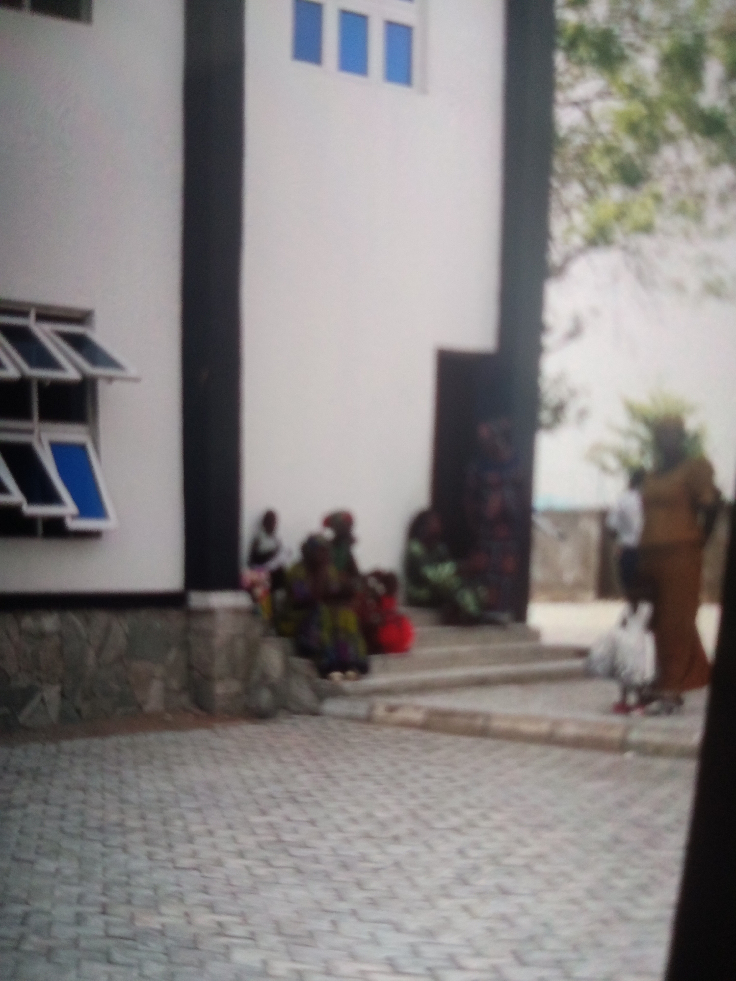 The image shows a blurred scene outside a building with a white facade. There are several windows on the building, some of which are open. In the foreground, a paved area is visible. In the middle ground, several people are gathered, some sitting on steps. One person is standing, wearing a brown garment. There are also some white objects, possibly birds or bags, on the ground. The overall lighting suggests it is daytime. The scene is likely located in Jalingo, Nigeria, given the provided context.
