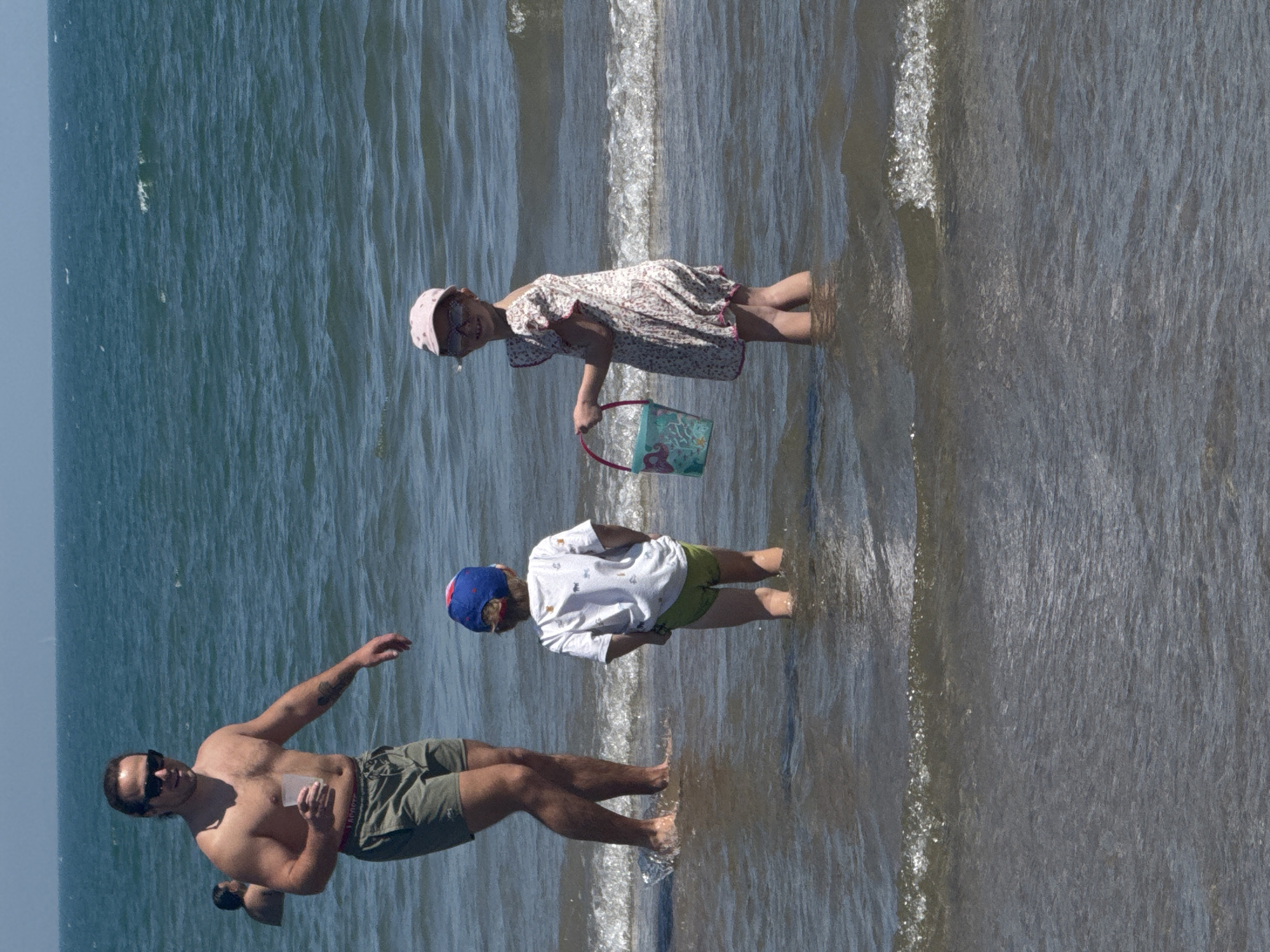 A father and two children are in the shallow water at the beach in Valencia, Spain. The father, shirtless with sunglasses and khaki shorts, is closest to the camera. A young boy wearing a white t-shirt, green shorts, and a blue and red hat stands further away from the camera. A young girl in a white sundress with pink hat and carrying a blue bucket stands farthest in the distance. All three individuals are standing in the water along the shoreline.