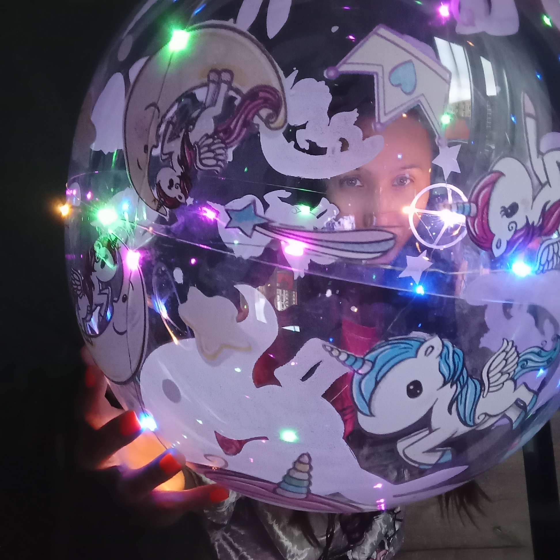 The image depicts a large, transparent, spherical balloon held by an individual. Inside the balloon, numerous small, multi-colored LED string lights illuminate, casting green, purple, blue, and orange points of light. The interior of the balloon also contains various cutouts or stickers, primarily featuring cartoon unicorns and related elements. One prominent unicorn, located in the lower right, has a blue mane, tail, and horn, with small wings. Another unicorn with a reddish-pink mane and wings is visible in the upper right. Other decorative elements include stylized stars, abstract shapes, a crown with a blue heart, and a small conical stack of rings.
A person's face, specifically their eyes and the bridge of their nose, is visibly reflected in the center of the balloon, looking directly forward. The person has dark eyes and eyebrows. Below the balloon, a pair of hands with bright orange manicured fingernails are seen holding the bottom of the balloon, with the left hand being more prominent. The background behind the balloon and person is largely dark and out of focus, providing minimal environmental detail. The image provides no visual information to determine the location in the city of Pervomaysky, Russia.
