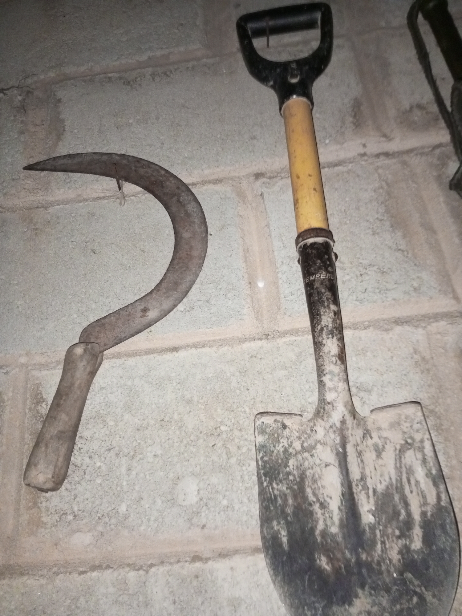 The image depicts a sickle and a shovel hanging on a concrete block wall. The sickle is on the left and the shovel is on the right. The shovel has a yellow handle and a black D-shaped grip. The image was taken indoors. There is no information about the city of Tinaquillo, Venezuela.