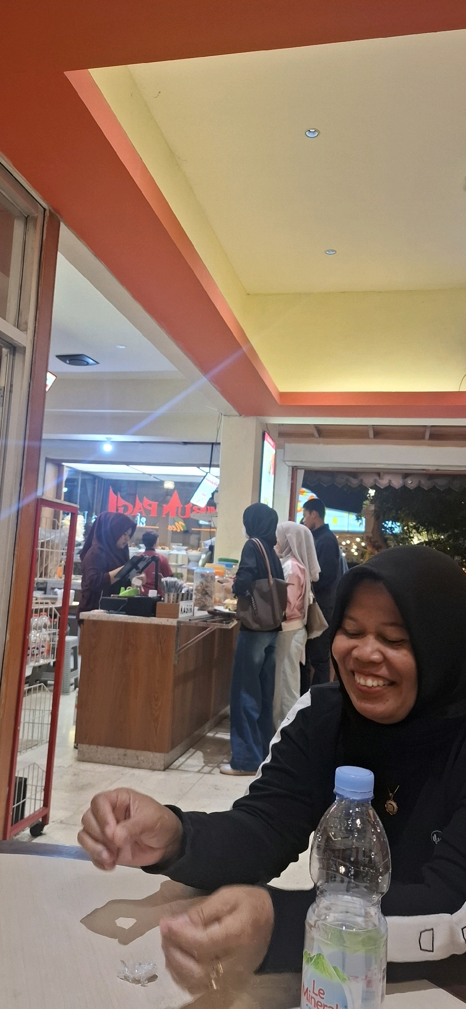 The image depicts an indoor scene, likely a food court or casual dining establishment, in Surakarta, Indonesia. In the foreground, a woman wearing a black hijab and a black top with white accents is smiling and looking to her right, out of frame. Her hands are resting on a light-colored surface, and a partially filled plastic water bottle with a blue cap is positioned to her right. A small, clear object, possibly ice or debris, is also on the surface in front of her.
In the mid-ground, several individuals are visible. A woman in a maroon hijab is standing behind a wooden counter, interacting with a device, possibly a cash register. To her right, another woman in a light-colored hijab and a pink top is standing. Further to the right, a woman in a black hijab and blue jeans is facing away from the camera, with a brown bag slung over her shoulder. A man in a dark shirt is standing behind them. The counter appears to be a food service station, with shelves and containers of food items visible. The background shows more of the establishment, with lighting and signage suggesting a commercial setting. The overall environment is brightly lit with artificial lights, and the ceiling features orange and yellow accents. The floor appears to be tiled.