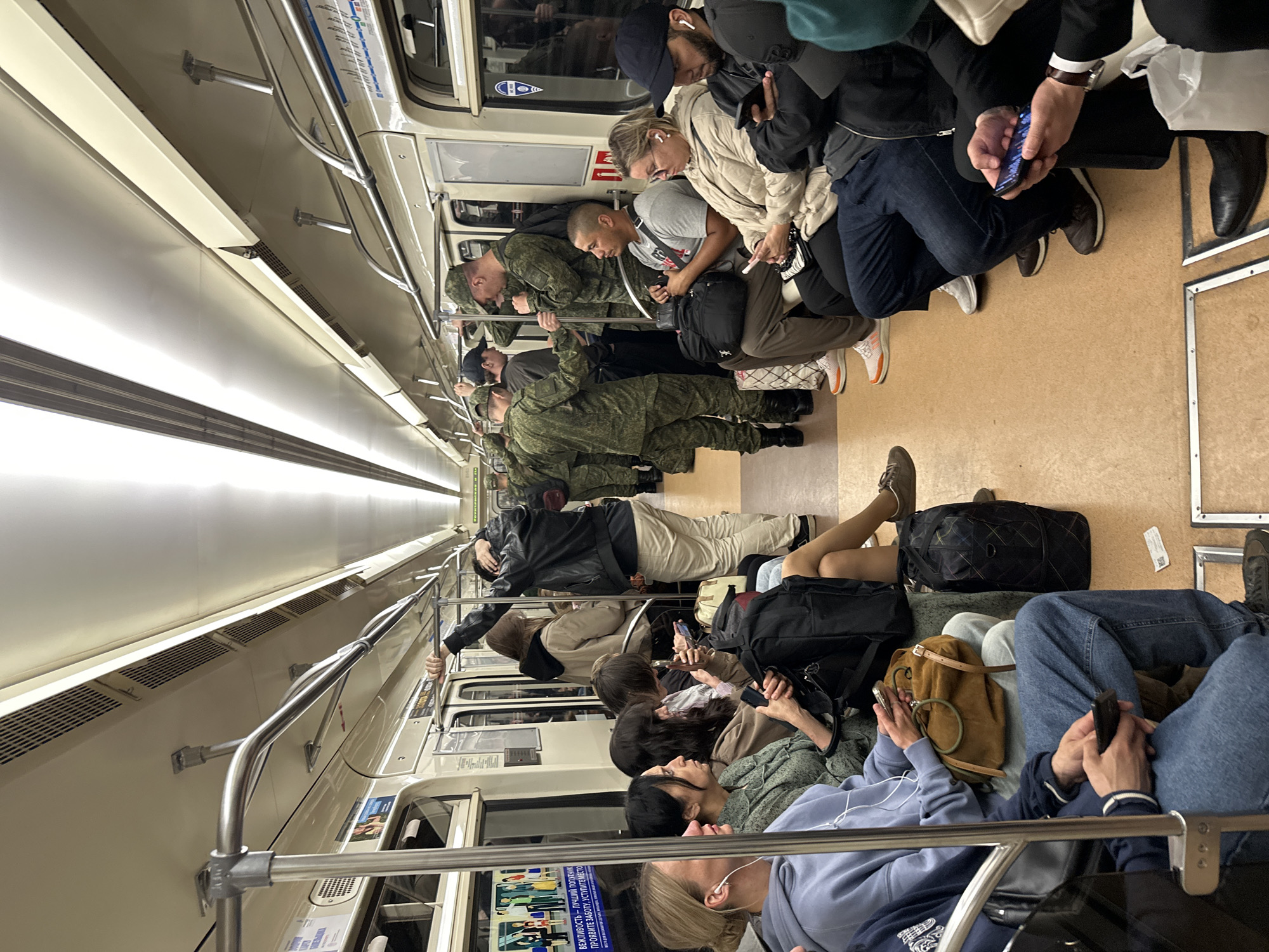 The image depicts the interior of a Saint Petersburg Metro train car during daylight hours, as indicated by the bright, artificial lighting. The car is moderately occupied with both civilians and military personnel. The flooring appears to be a light brown, worn material, and the walls are a cream color, featuring various advertisements and informational posters. Handrails, made of polished metal, run along the ceiling and sides of the car for passengers to hold onto.
In the foreground, several civilians are seated. On the left, a woman in a light blue hoodie and earphones is looking at her phone. Next to her, a woman with dark hair in a green patterned shirt is also looking at her phone. Further down the row, other civilians are seated, some engaged with their phones, others looking forward or around the car.
The middle and back sections of the car show a number of individuals in military uniforms, primarily of a camouflage pattern. Some are standing and holding onto the handrails, while others are seated. Their presence suggests a potential military mobilization or deployment. One man in a military uniform is interacting with a civilian man who is seated.
Luggage, including backpacks and bags, is visible throughout the car, placed on the floor or on the seats, indicating that passengers are traveling. The overall atmosphere appears to be one of routine travel, with the presence of military personnel adding a notable element to the scene.