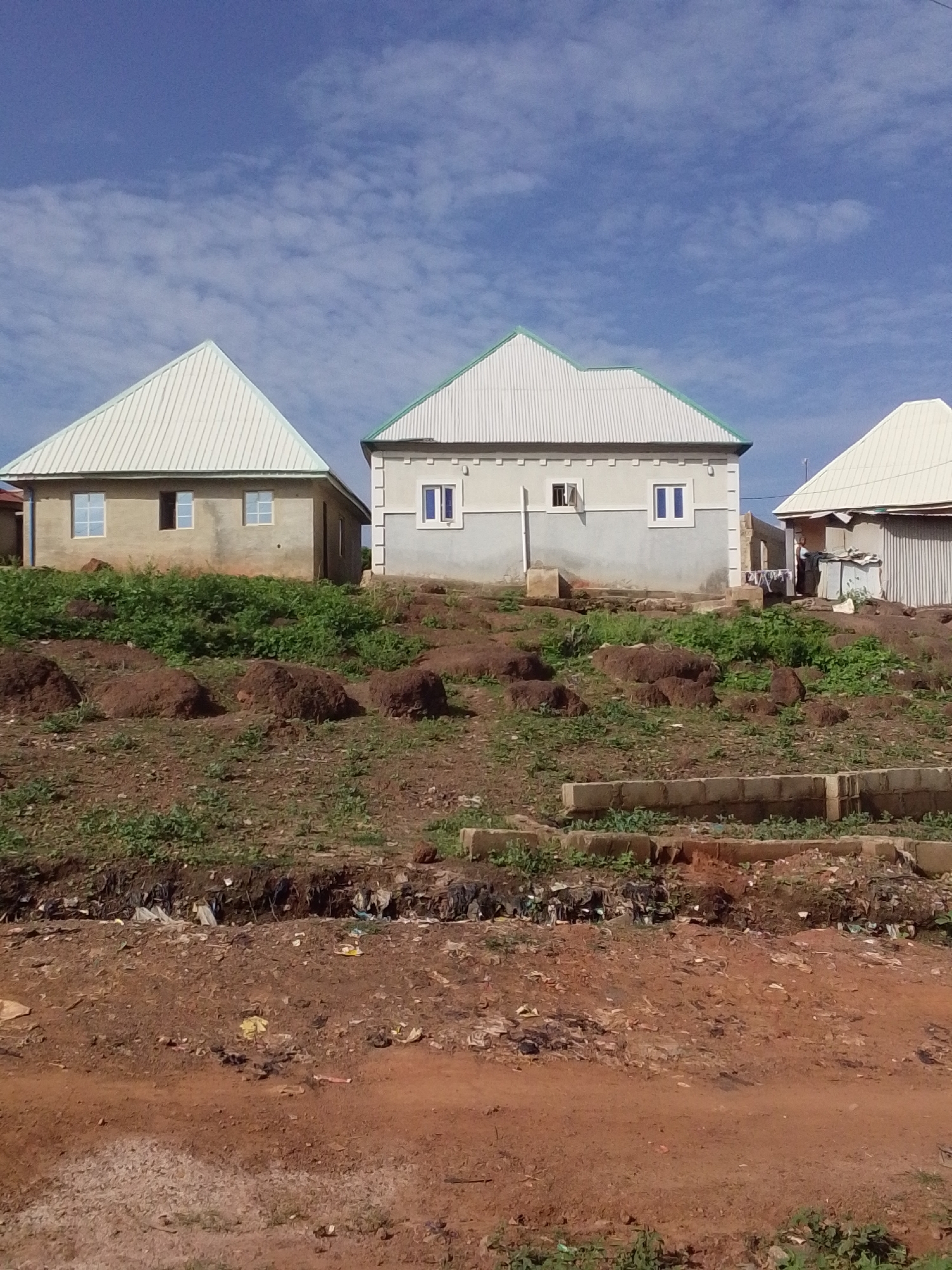 This image depicts a residential scene in Keffi, Nigeria, captured on a clear day with a bright blue sky and scattered white cirrus clouds.
In the background, three single-story houses are visible, situated on an elevated, uneven terrain. From left to right:
The leftmost house is unfinished, featuring unpainted light brown concrete block walls and a corrugated metallic roof with a light green trim. It has four rectangular window openings; two contain glass panes, one is an open dark void, and one appears to be boarded or dark.
The central house is completed, displaying light grey painted walls with decorative darker grey L-shaped trim elements. It has a similar corrugated metallic roof with a light green trim. Several windows with dark blue frames are present, some appearing closed, others slightly open. A small white garment is hanging near the far-right window.
The rightmost house is partially visible, appearing to have corrugated metallic walls and a light-colored corrugated metallic roof. A figure, consistent with a person wearing a light-colored top, is partially discernible near its entrance. Laundry is visible hanging to dry near this structure.
The midground consists of reddish-brown soil with sparse green vegetation and several large, irregularly shaped mounds of dark brown earth.
The foreground features an unpaved reddish-brown dirt path or road. A distinct horizontal trench or depression runs across this foreground section, filled with a significant accumulation of refuse, including plastic bags, wrappers, and general debris. Concrete blocks are visible in the lower foreground, possibly forming part of a low wall or foundation. The ground is uneven and shows signs of erosion and human activity.