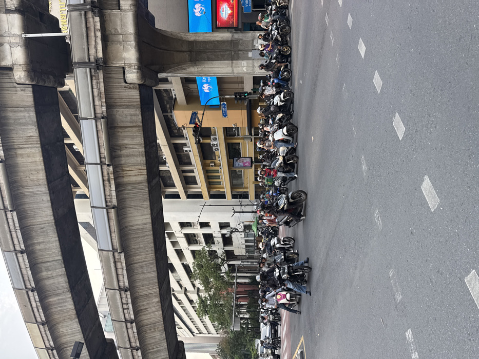 The scene depicts a busy street intersection in Bangkok, Thailand, identified by the visible "Krungthai" bank signage and the Thai text "ราชดำริ" (Ratchadamri) on a blue street sign, locating it at Ratchadamri Road, likely near the Ratchadamri BTS Skytrain station. Elevated concrete structures, characteristic of the BTS Skytrain tracks, span the upper portion of the image. Below, approximately 30 to 40 individuals are mounted on various motorcycles and scooters, stationary at a red traffic light, forming dense rows across multiple lanes on an asphalt road. The majority of riders wear helmets, and some are observed wearing face masks. In the background, multi-story buildings include a yellow-facade structure with visible air conditioning units and a larger, lighter-colored corporate building. A traffic light displaying a red signal is positioned on the right side of the intersection. Minimal vegetation is visible to the left of the road.