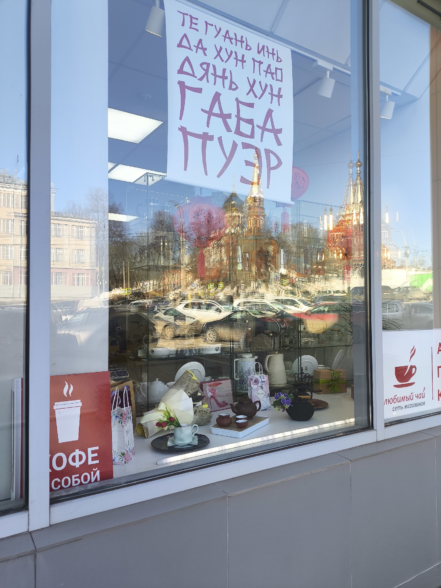 The image depicts a street-level shop window in Izhevsk, Russia, displaying merchandise primarily related to tea and coffee. Inside the window, a curated arrangement of white and dark-colored teapots, cups, saucers, pitchers, and gift items is visible. Notable display elements include a light blue teacup holding red tulips and small purple flowers near a dark teapot, and a wrapped gift box tied with a green ribbon.
Affixed to the interior of the window are several signs: a red vertical sign on the left features a white coffee cup icon and the Russian text "КОФЕ С СОБОЙ" (Coffee to Go). On the right, a red horizontal sign displays a red teacup icon and the Russian text "ЛЮБИМЫЙ ЧАЙ сеть магазинов" (Favorite Tea, chain of stores). A large white banner hanging from the ceiling, partially visible in the upper center, has dark pink/purple Russian text listing types of Chinese tea: "ТЕ ГУАНЬ ИНЬ ДА ХУН ПАО ДЯНЬ ХУН ГАБА ПУЭР".
The exterior environment is extensively reflected in the window's glass. The most prominent reflection is that of the Saint Michael's Cathedral (Mikhailovsky Cathedral) in Izhevsk, identifiable by its numerous golden onion domes and red brick walls, situated across a street. The reflection also shows a street with numerous parked and stationary vehicles, including sedans and SUVs of various colors (white, red, dark), other multi-story city buildings, and bare trees. Yellow pedestrian crossing signs are faintly discernible in the reflections. The sky appears clear and bright, indicating daylight conditions. No people are visible in the scene, either inside the shop or in the reflections.