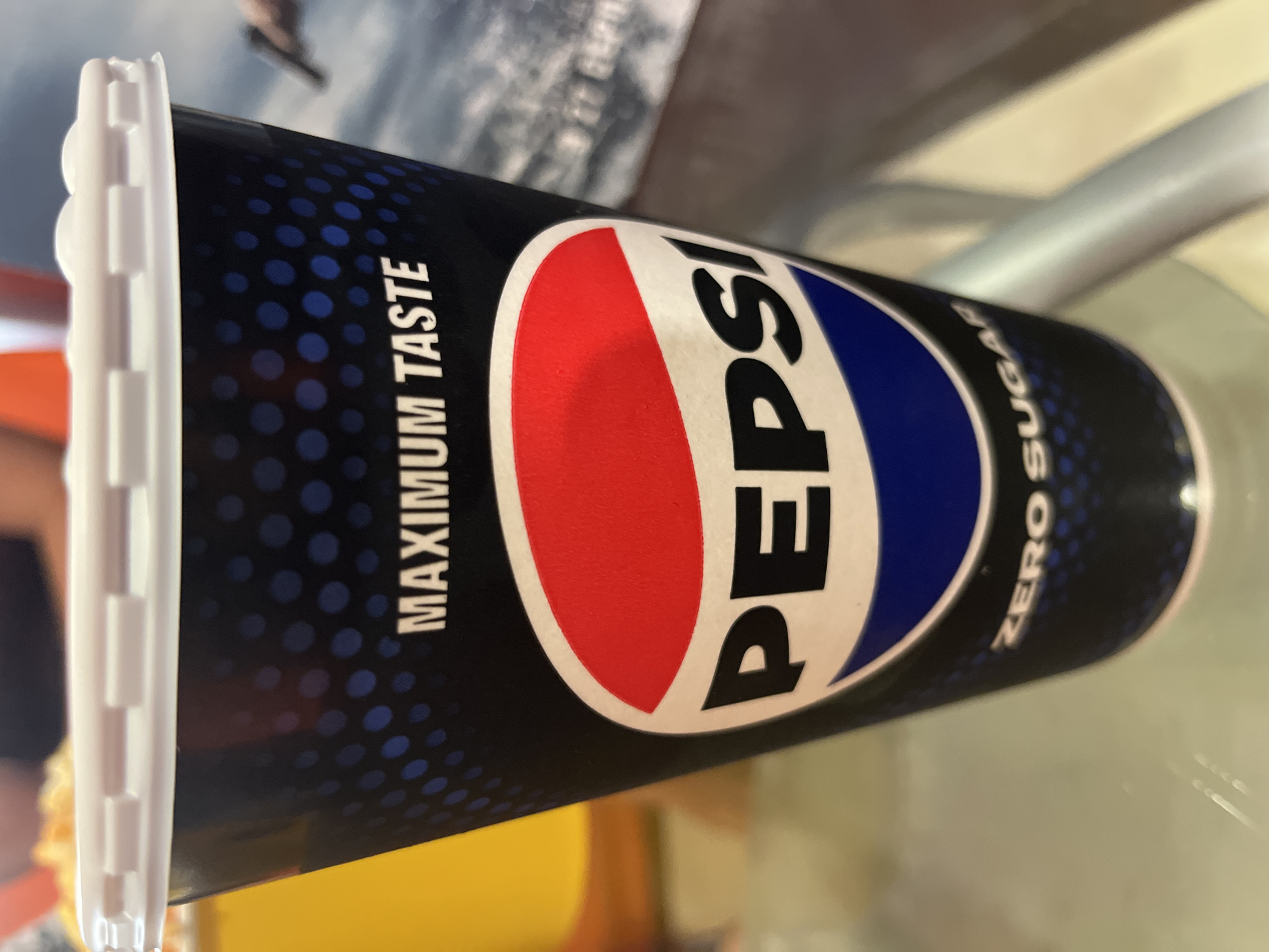 A close-up, eye-level, vertical shot shows a Pepsi Zero Sugar cup, with a white lid, positioned on a glass table. The cup has a black background with a pattern of blue dots and text that reads "MAXIMUM TASTE" above the iconic Pepsi logo, which is a red, white, and blue circle. Below the logo, the text "ZERO SUGAR" is visible. The background is blurred, showing a yellow object on the left and a muted, out-of-focus image on the right, possibly a poster or screen. The lighting appears to be natural, with soft highlights on the cup and table. No specific location in Kharkiv, Ukraine, is identifiable from the image.