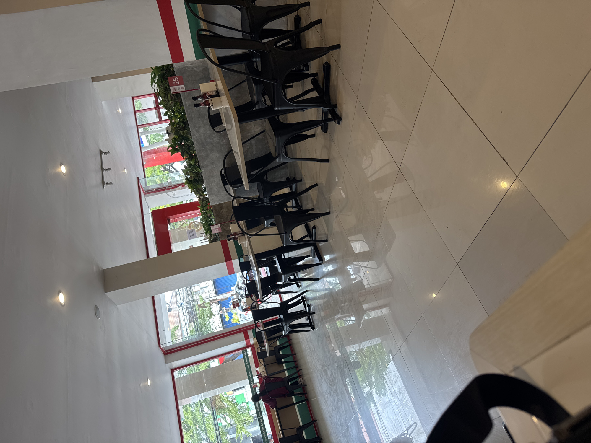 This is an interior shot of a restaurant located in Denpasar, Indonesia. The restaurant features a modern, minimalist design with a lot of natural light coming in from the large windows. The flooring is made of large, beige tiles that reflect the light, creating a bright and airy atmosphere. The tables are light-colored and have black metal legs. They are set with black metal chairs that have a simple, industrial design.
In the background, there are several plants, suggesting an attempt to bring some greenery indoors. There are also some bottles on a table, possibly condiments, and a sign with the number "25" on it, likely indicating a table number or a promotional offer. A person is seated at a table by the window, looking away from the camera. The overall impression is a clean, well-lit, and somewhat empty restaurant space.
The exterior view through the windows shows trees and buildings, hinting at an urban environment within Denpasar. The red and white striped accents on the exterior of the building are a prominent visual feature.