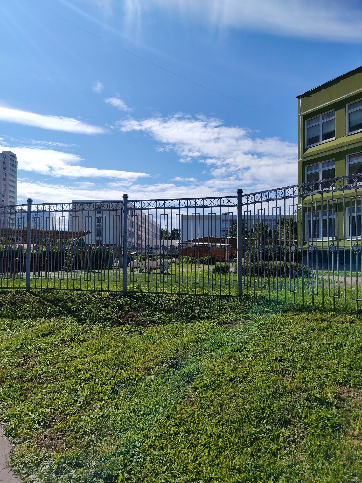 A sunny day in Zelenograd, Russia, features a metal fence in the foreground, separating a patch of green grass from a playground and apartment buildings in the background. The fence has a decorative pattern of circles and vertical rectangular elements. Behind the fence, a playground area is visible with some structures, including a slide and what appears to be a small canopy. Further in the background, several multi-story apartment buildings, characteristic of Soviet-era architecture, rise against a bright blue sky dotted with white clouds. One building on the right is painted a light green. The overall scene depicts a typical urban environment on a clear day.