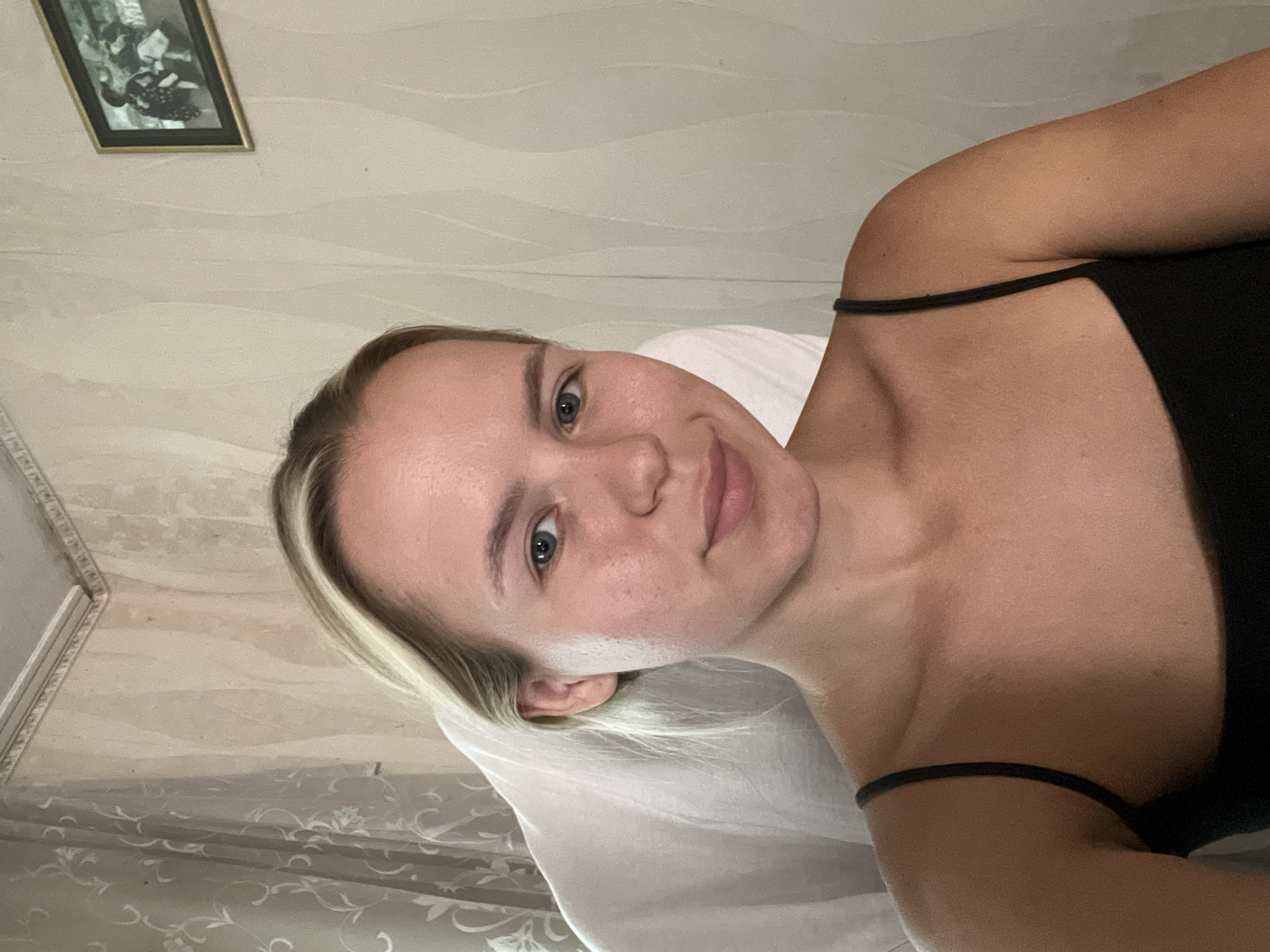 A young woman with blonde hair and blue eyes is looking at the camera. She is wearing a black tank top. She is sitting in a chair with a white cover. The background is a wall with a patterned wallpaper. There is a framed picture on the wall to the right.