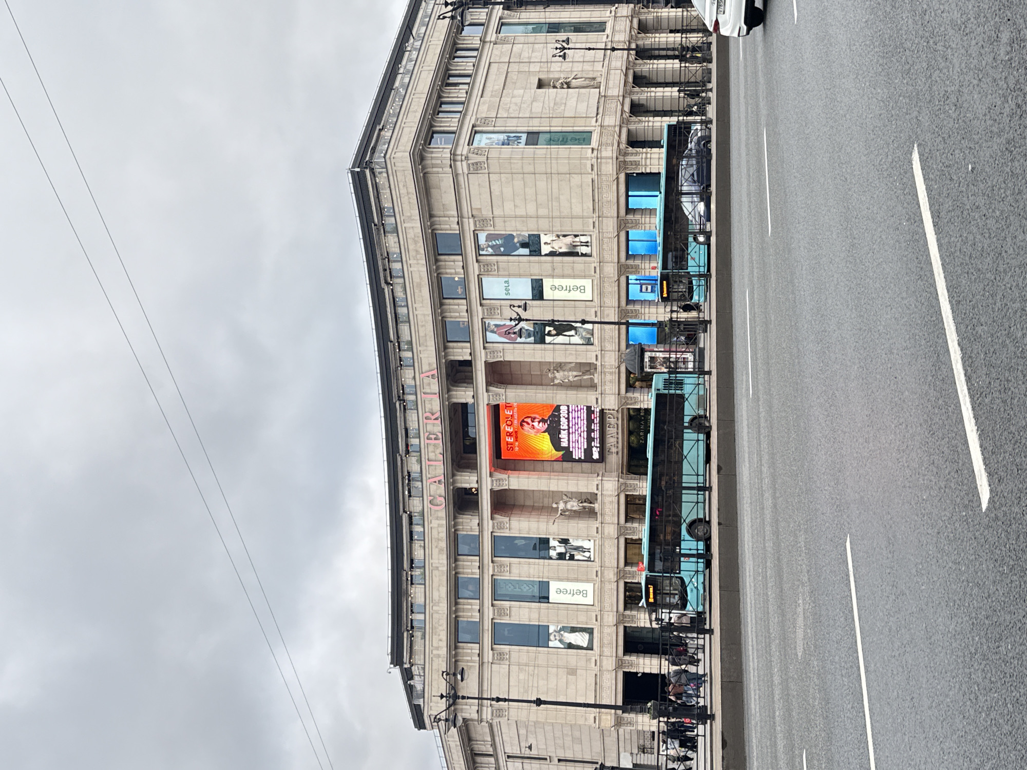 Here is a description of the image:
The image depicts a street scene in Saint Petersburg, Russia. The most prominent feature is the "GALLERIA" shopping mall, its name emblazoned vertically on the building's facade. The building's architectural style includes classical elements with pillars and decorative elements. A large digital billboard is visible on the building displaying an advertisement featuring a person and some text. Below the billboard are displays with posters of "BeFree".
A blue bus is parked on the street next to the shopping mall. The bus has tinted windows and the name of the bus route is visible on it, stating "ZAPINA 9 ROD..." Some people appear to be near the bus stop.
A wide asphalt road is in the foreground with white lane markings, indicating a multi-lane traffic way. Overhead electrical or communication lines are visible against a cloudy sky.