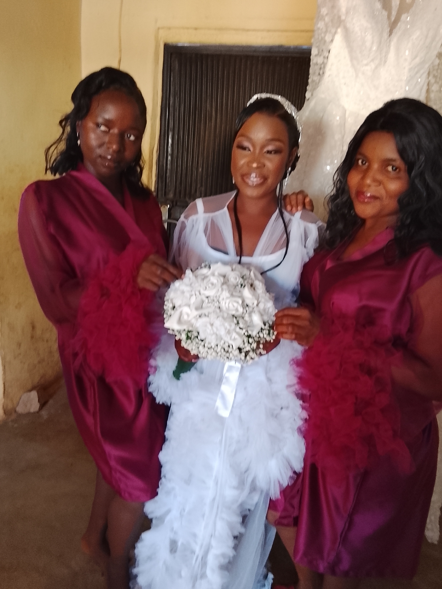 Three women pose for a photo indoors. The woman in the center wears a white dress and a tiara, holding a bouquet of white flowers. The other two women are on either side of her, wearing maroon robes with feathered trim. The background includes a window with closed blinds and a white garment hanging. The location is Unknown, Unknown.