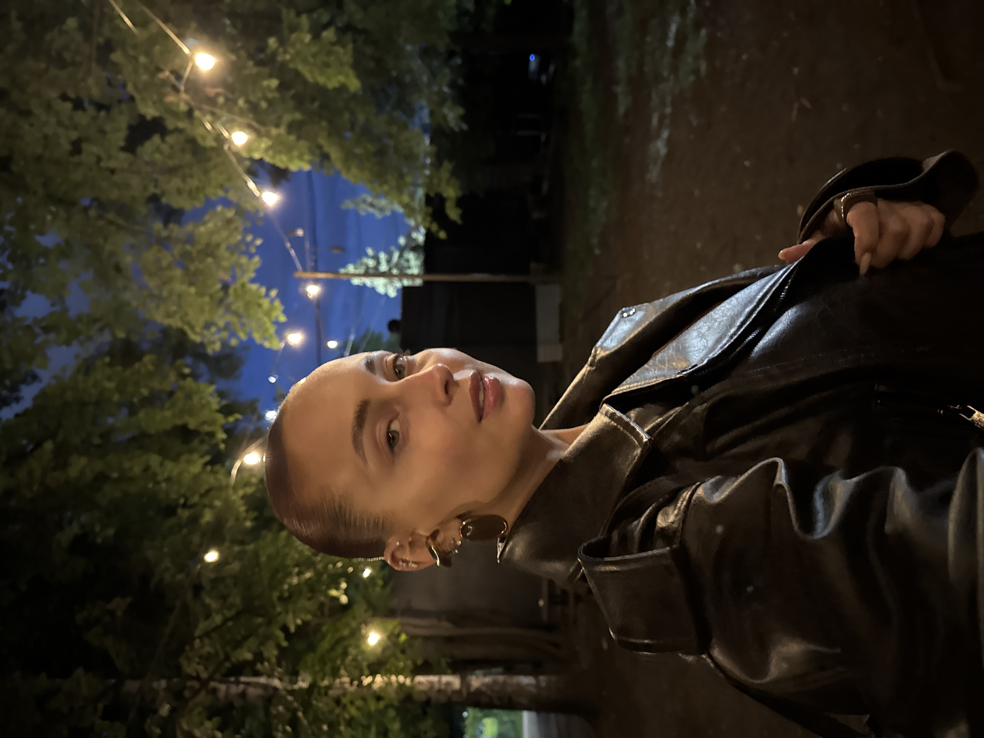 The image features a close-up portrait of a young woman outdoors at night in Izhevsk, Russia. She is positioned on the right side of the frame, looking directly towards the camera with a calm and composed expression. She has light skin, dark hair pulled back, and is wearing subtle makeup, including a natural lip color. Notable accessories include prominent, possibly gold-toned, abstract-shaped earrings.
The woman is wearing a dark, likely black or deep brown, leather jacket with visible textures and folds, and a zippered front. Her right hand, adorned with a ring and manicured nails, is visible resting on her jacket.
The background is an outdoor setting at night, illuminated by warm string lights strung between trees with green foliage. The sky is a dark blue hue, indicating either late twilight or nighttime. The ground appears dark, possibly unpaved earth or a dimly lit path. The scene suggests a relaxed evening ambiance, perhaps at an outdoor cafe, garden, or event. The woman is posing for the photograph. No visible text is present in the image.