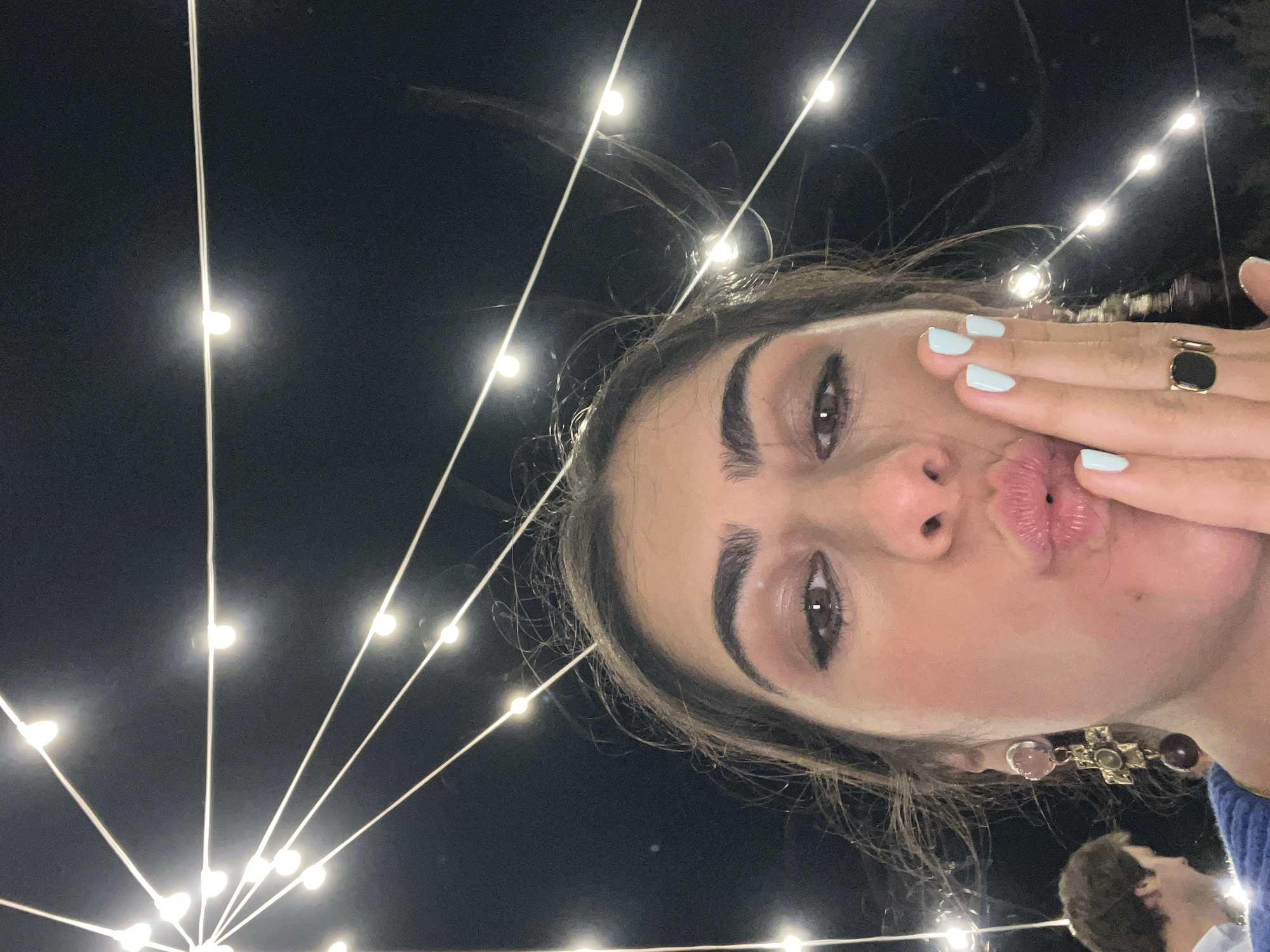 The image captures a close-up, low-angle selfie of a woman with dark hair and full lips, puckering her mouth and resting her right hand on her cheek. She is wearing a blue top, a large cross-shaped earring with colorful stones, and a gold ring with a black square stone on her right index finger. Her fingernails are painted a light blue. In the background, strings of small, bright lights are strung against a dark night sky, forming a radial pattern from the top left corner of the image. To the left and out of focus, a man in a light-colored shirt is partially visible. The scene appears to be outdoors at night, possibly in the city of Fasano, Italy, given the context.