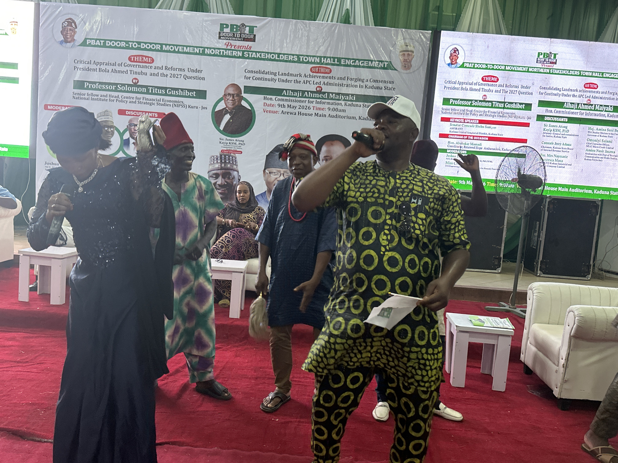 Here is a description of the image:
The image captures a political or community event, likely taking place in Arewa House Main Auditorium, Kaduna State, as indicated on a backdrop banner. The event seems to be a "PBAT Door-to-Door Movement Northern Stakeholders Town Hall Engagement," focused on discussing governance and reforms under President Bola Ahmed Tinubu.
Several people are visible in the frame. In the foreground, a man in a patterned shirt and cap stands, speaking into a microphone and holding a small flag or paper. To his left is a woman in a black dress also holding a microphone. Behind them, other individuals are visible, dressed in traditional or formal attire.
The backdrop banner features the event title, theme, sub-theme, and names of speakers and discussants. The banner suggests the event involves discussions around "Consolidating Landmark Achievements" and "Forging a Consensus" for continuity under the ruling political party (APC).
The scene is on a red carpet. There are small white tables, a white chair, and a fan in the background.