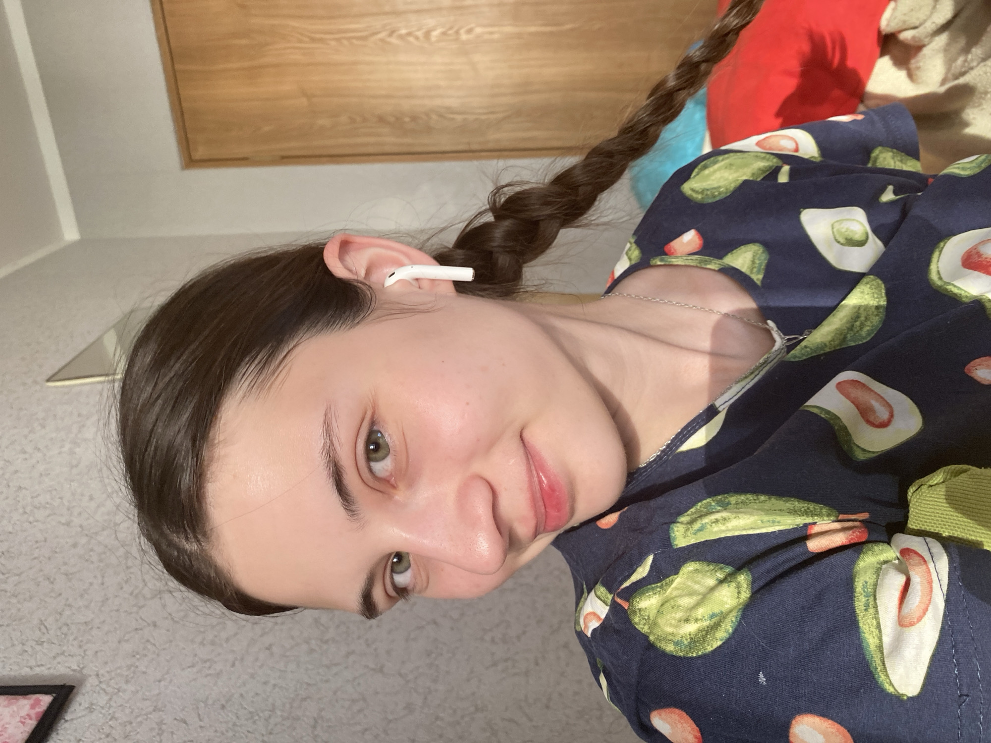 A close-up portrait shows a young woman indoors. She has brown hair styled in a braid, fair skin, and is wearing an earbud in her left ear. She has a necklace and wears a navy blue garment with an avocado pattern. A wooden surface and a light-colored wall are visible in the background. There is no indication of a location in Votkinsk, Russia.