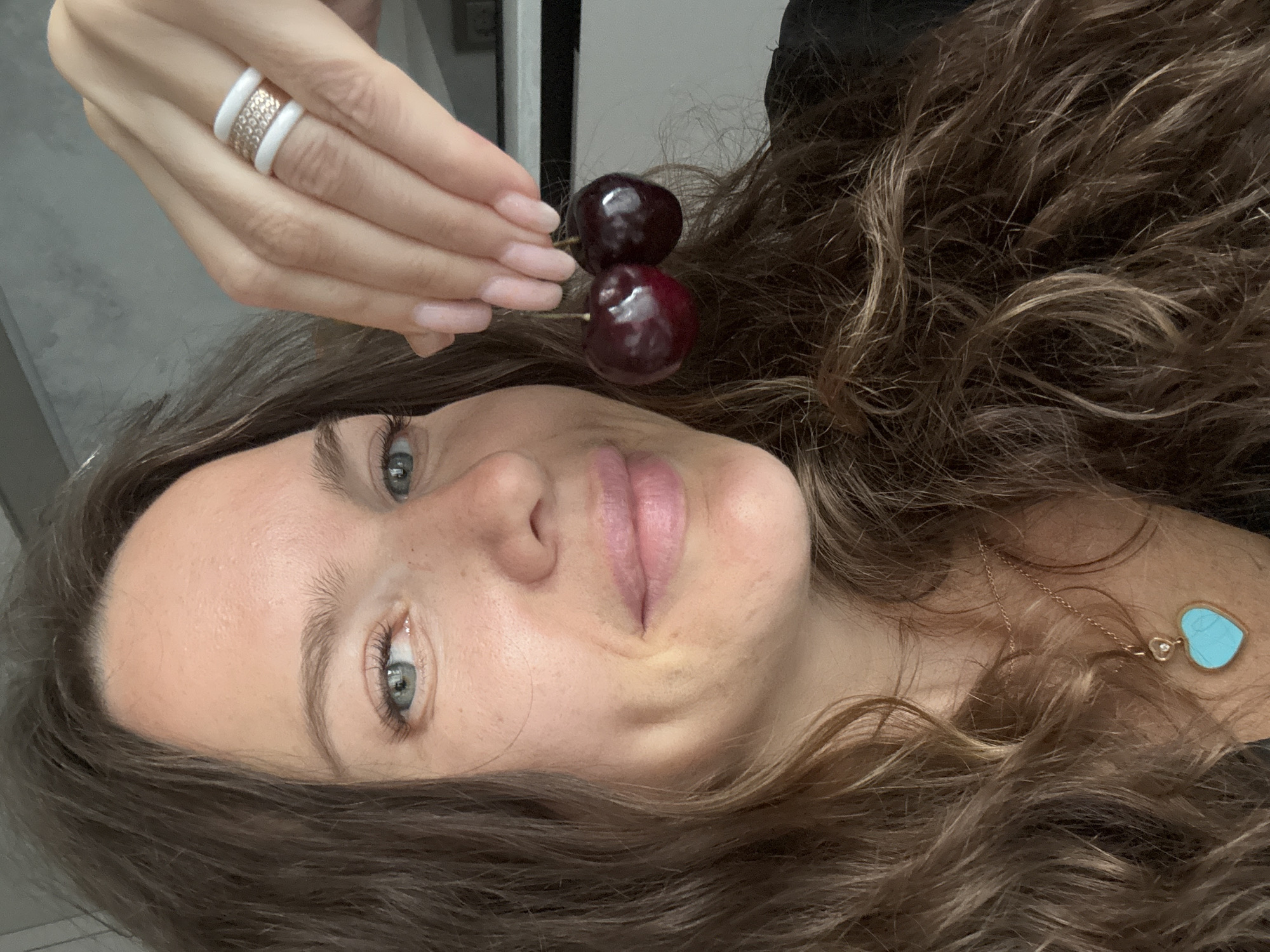 A woman, with brown hair and blue eyes, is taking a selfie. She is holding two cherries between her thumb and index finger of her right hand, which is adorned with a white and rose gold ring. The woman is also wearing a turquoise heart-shaped pendant necklace. Her lips are slightly parted, and she has dimples on her cheeks. She is wearing a black top. The background is blurred, suggesting an indoor setting.