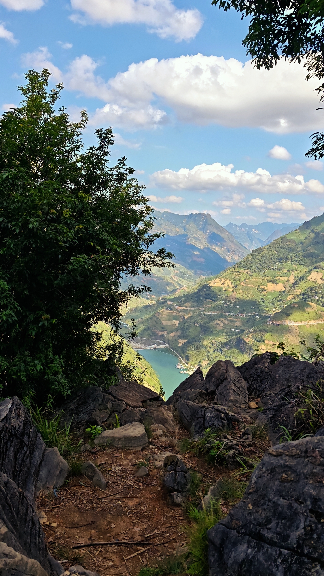 A scenic view of the mountainous region of Đồng Văn Commune, Vietnam. In the foreground, a rocky outcrop, featuring jagged, dark gray stones, leads to a narrow, earthy trail. Patches of green vegetation punctuate the rocks and trail. To the left, a dense tree with lush green foliage partially obscures the view. Beyond the rocks, a wide valley unfolds, showcasing layers of rolling hills and rugged mountains. The hills are covered in verdant vegetation, interspersed with roads and small settlements. In the distance, mountain peaks rise toward a blue sky dotted with fluffy white clouds. A body of water, reflecting a turquoise hue, lies within the valley, with visible structures along its banks. The image is shot from a high vantage point, looking down into the valley, creating a sense of depth and scale.