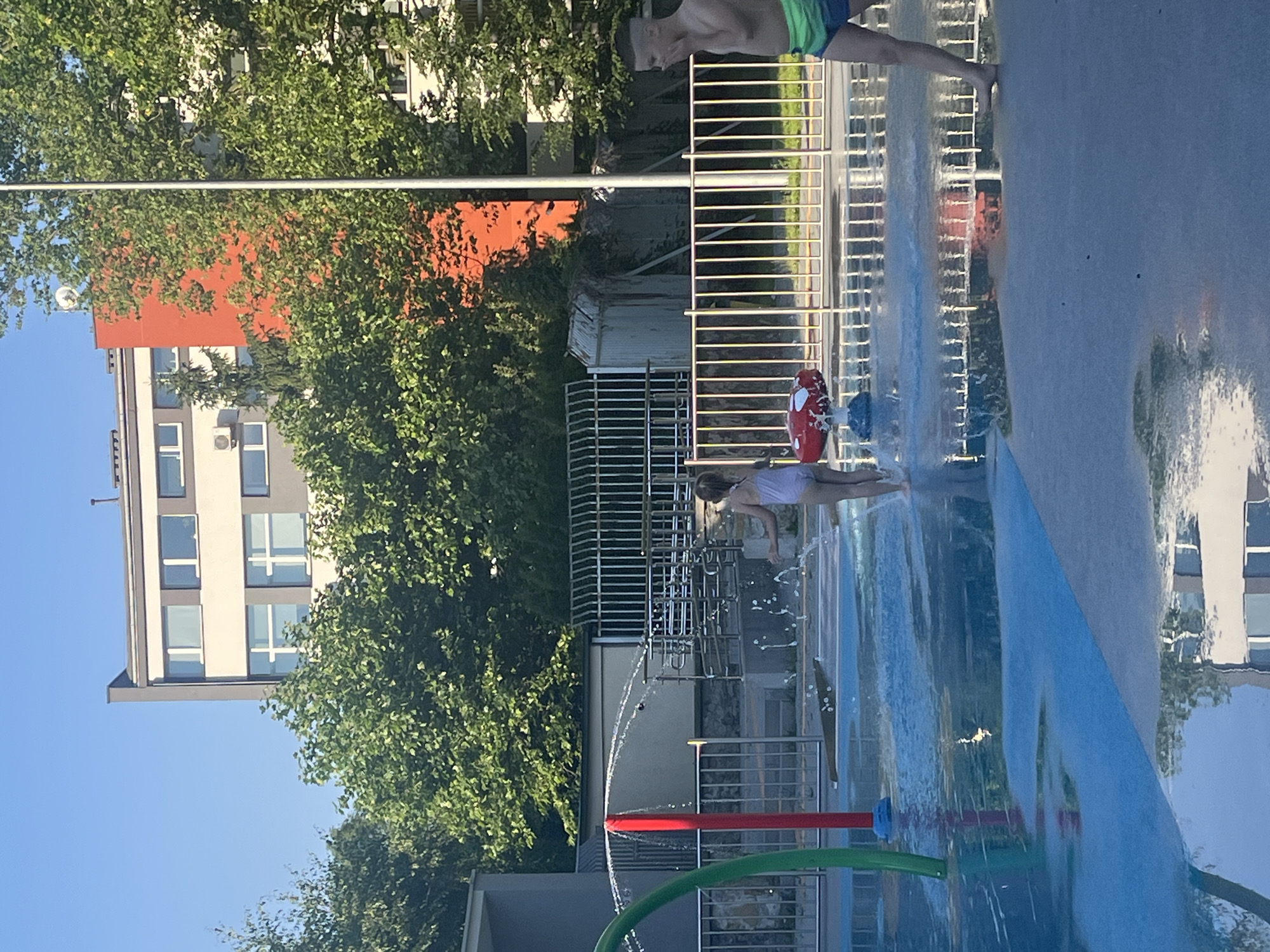 In an outdoor splash pad area, two children, a girl and a boy, are visible. The girl, wearing a white swimsuit, stands with her back to the camera, facing a water feature that is spraying water. A red mushroom-shaped toy is also visible near her. The boy, on the right side of the image, is wearing blue and green swim trunks and is facing towards the left, with his right hand covering his mouth. The splash pad has blue and grey surfaces, with shallow water covering parts of it, reflecting the surrounding environment. In the background, there are metal fences, lush green trees, and multi-story buildings. One building has an orange facade, and another is light-colored with dark-trimmed windows. The sky is clear and blue.