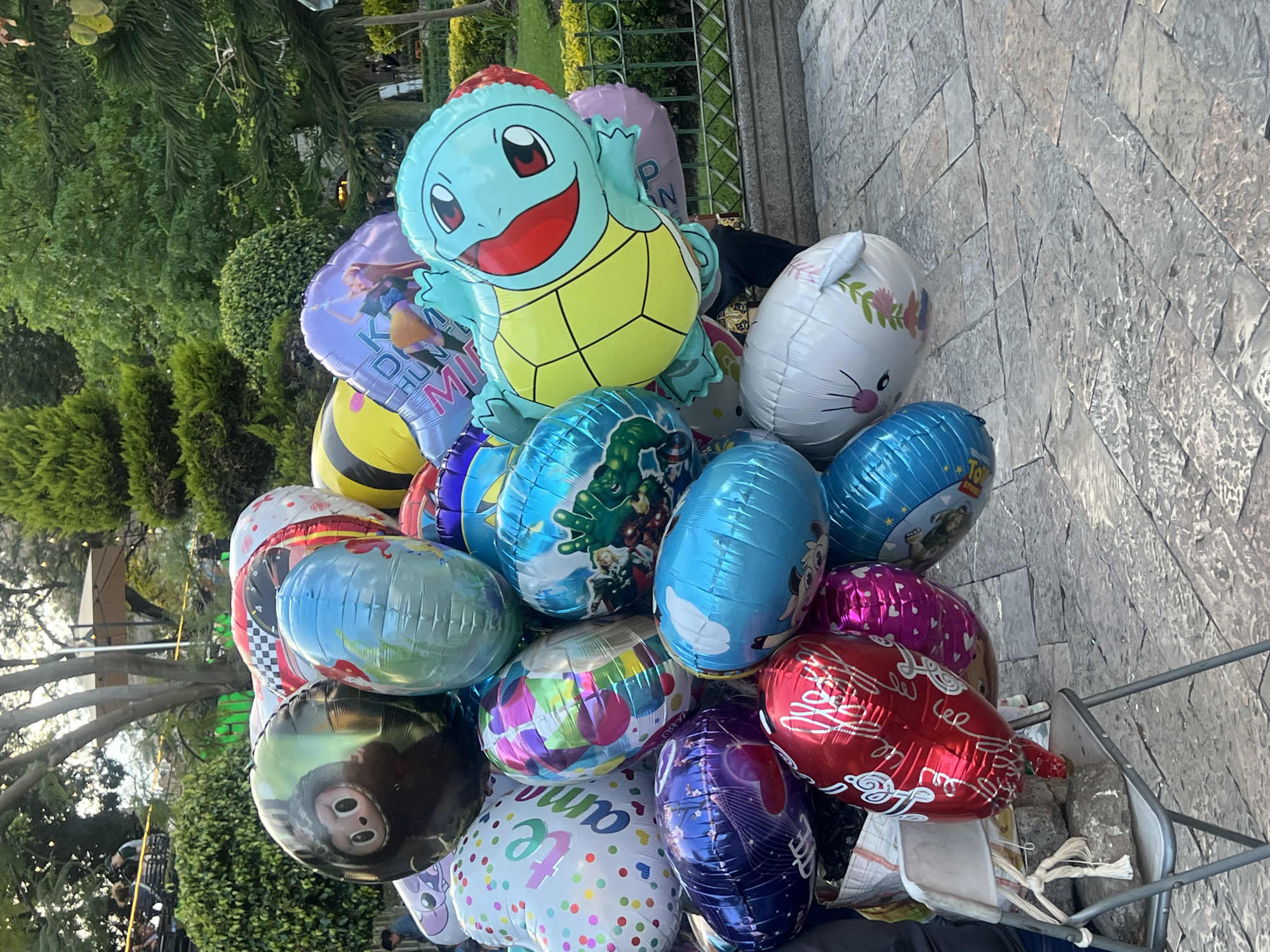 A medium shot of a collection of balloons in an outside location. There is a large cluster of balloons with different shapes and cartoon characters. A Squirtle balloon sits at the top of the cluster. The balloons are sitting on top of a cart on a stone-paved walkway. In the background are green trees and a metal fence.
