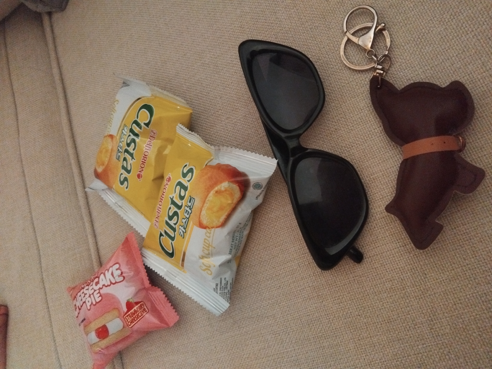 On a light beige textured surface, likely fabric, there is an arrangement of personal items and snacks. Two packages of "Custas" soft cup cakes, with yellow and white packaging, are stacked on top of each other. Below them is a small, pink package of "Cheesecake Pie". To the right, a pair of black sunglasses is placed next to a brown leather dog-shaped keychain with a silver keyring.