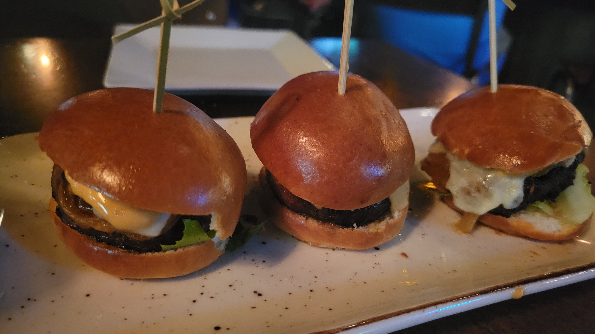 The image shows three mini burgers, or sliders, arranged in a row on a speckled white rectangular plate. Each slider has a golden-brown bun, a dark patty, and appears to have some sort of sauce or cheese. The leftmost slider has lettuce and what looks like caramelized onions visible. The middle slider has a light-colored sauce. The rightmost slider has melted white cheese dripping down the sides. Each slider is secured with a wooden skewer. The plate is set on a dark, possibly wooden, surface. The background is blurred, with hints of ambient lighting and out-of-focus objects, suggesting an indoor setting like a restaurant. There are no people or animals visible in the image, and no specific location in Prévost, Canada, is identifiable from the visual information provided.