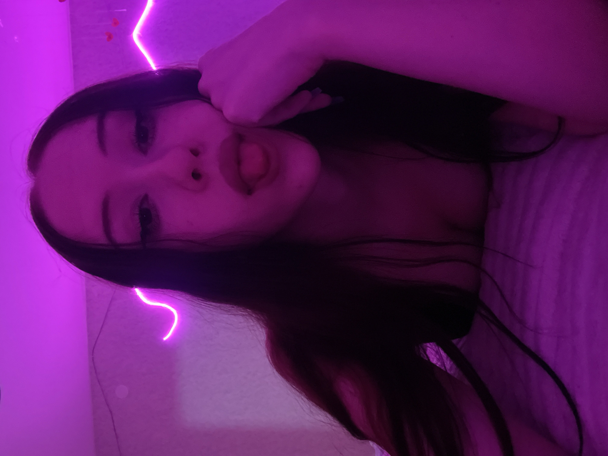 A young woman with long dark hair is lying down in a bed. She is wearing a dark top that reveals her shoulders and chest. Her face is tilted upwards and her tongue is sticking out. She is wearing makeup. The room is lit with purple neon lights. One light forms a wavy line along the wall behind her. Another light is a heart shape. There is a pillow or blanket under her head. The room appears to be a bedroom. The image does not contain any information that could be used to determine the location in the city of Votkinsk, Russia.
