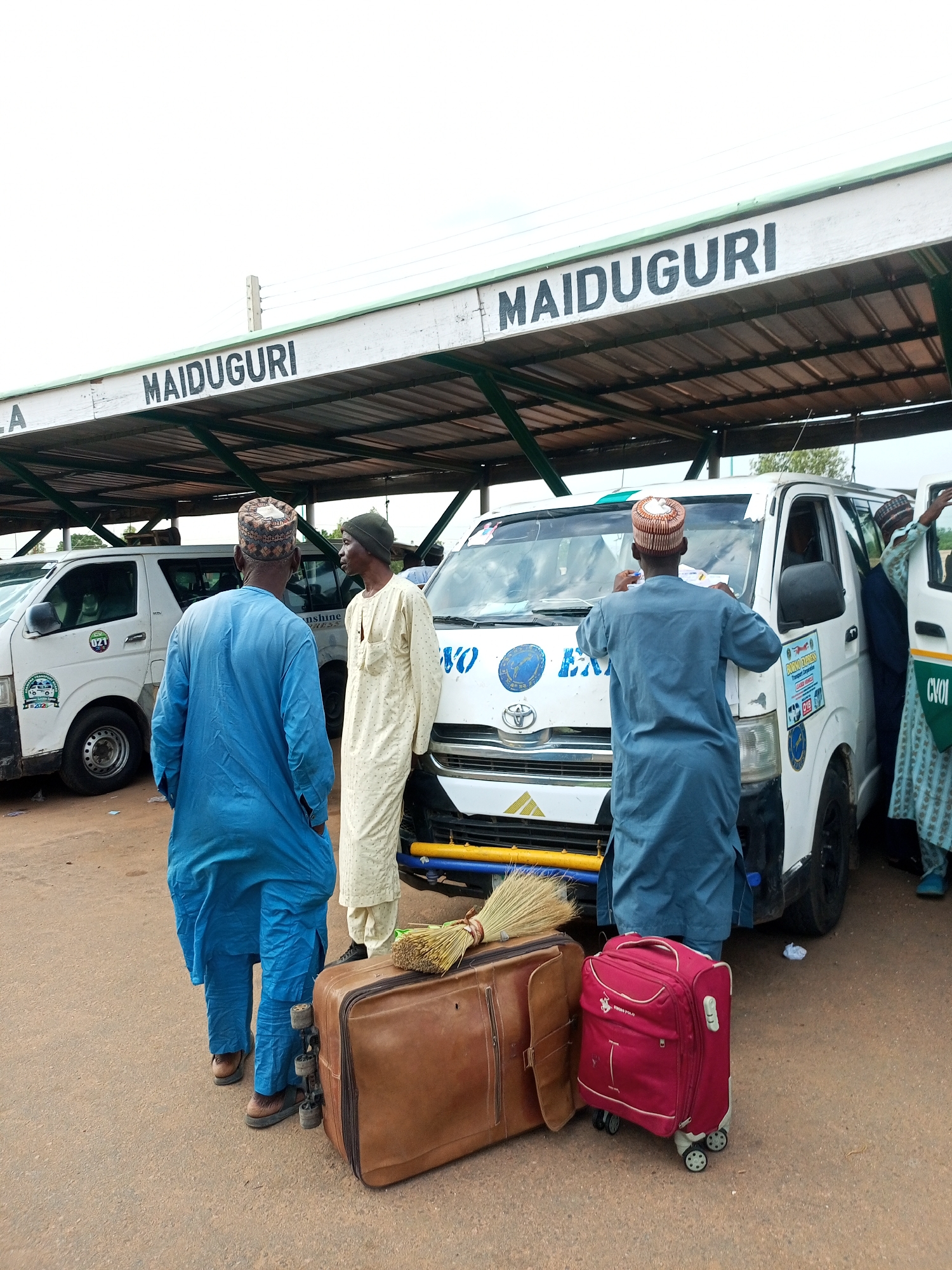 This image depicts a scene at what appears to be a transportation hub in Gombe, Nigeria. Several individuals, dressed in traditional Nigerian attire, are gathered around a white minivan. Two men are standing, one in a blue robe and the other in a cream-colored robe. A third man in a blue robe is leaning over the hood of the minivan, seemingly attending to something on the vehicle. Luggage, including a large brown suitcase with wheels and a red suitcase, is placed on the ground next to the minivan, suggesting travel. A broom is also visible resting on the brown suitcase.
The setting includes a covered structure overhead, with the word "MAIDUGURI" painted in large black letters on a white background, indicating a destination or route. Another white minivan is parked to the left, and part of a third minivan with "CVOI" visible on its side is on the right. The ground appears to be paved with dirt or gravel. The lighting suggests it is daytime, with overcast skies. The overall atmosphere seems to be one of preparation or departure for a journey.