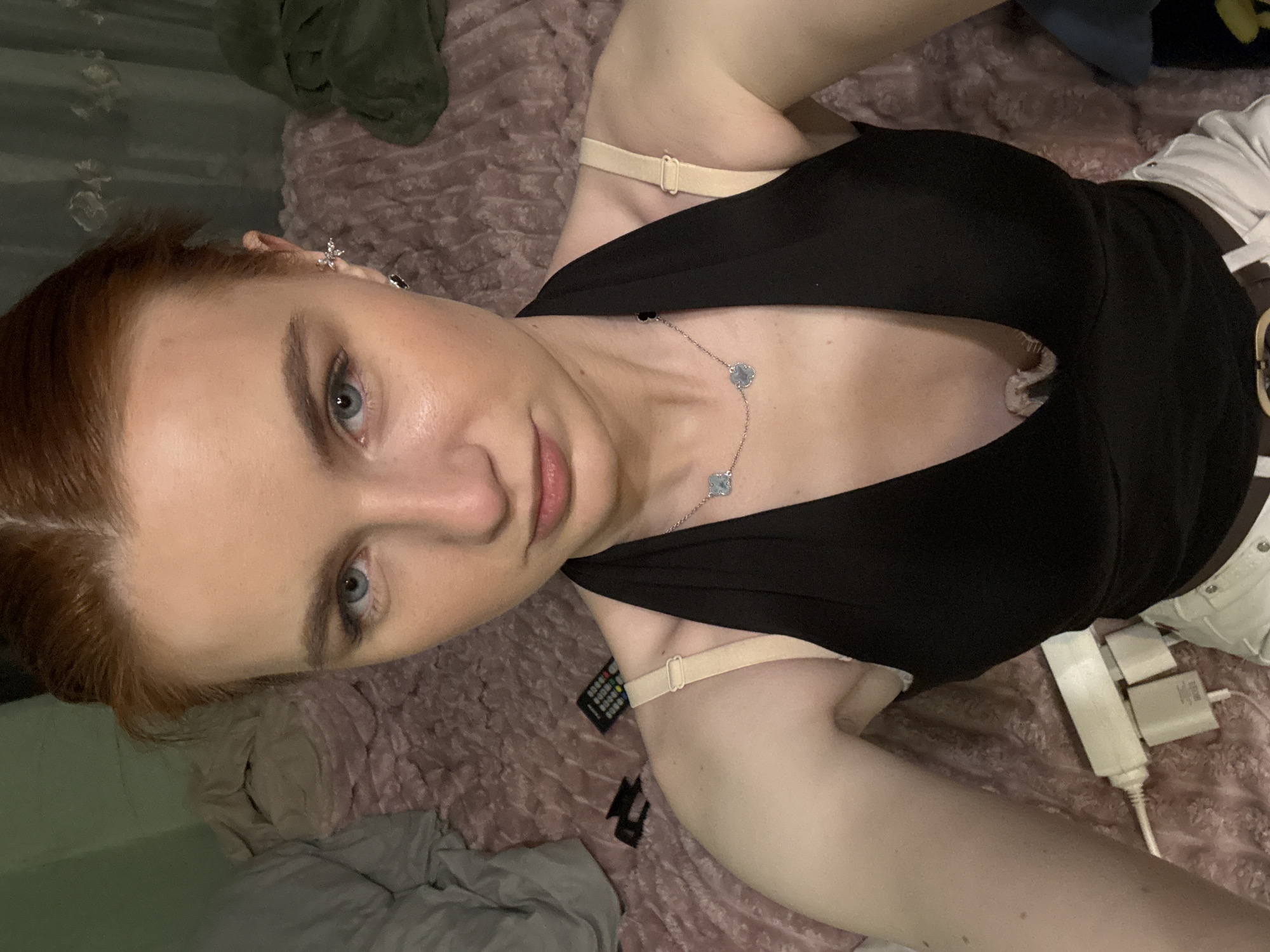 A woman with light skin, red hair pulled back, and blue eyes is lying on a pink textured blanket, wearing a low-cut black top and white pants with a brown belt. A silver necklace with flower-shaped pendants is visible on her chest. An earring is visible on the left side of her face. A remote control, charging cables, and plugs are scattered on the blanket. The location cannot be determined from the image.