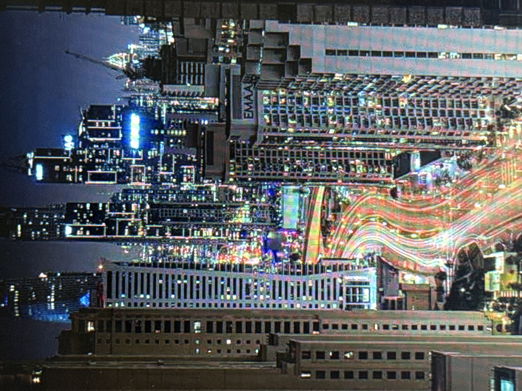 This media file captures an abstract, glitchy, night-time aerial view of a city. The dominant subjects are a dense collection of illuminated buildings and structures, appearing almost like a futuristic or digital cityscape due to the visual artifacts. There are no discernible people or specific objects like vehicles or street furniture. The scene is characterized by vibrant, chaotic light trails and illuminated windows, suggesting a busy urban environment.
The image is highly stylized and abstract, making it difficult to pinpoint a specific activity or event. The long exposure light trails of what appear to be roadways or waterways contribute to a sense of movement and energy. The time of day is clearly night, evidenced by the extensive artificial lighting and dark sky, though the darkness is heavily obscured by the bright lights and visual noise. The weather is not discernible.
Notable details include the fragmented and distorted nature of the image, with blocks of pixels appearing out of place or overly saturated. The color palette is dominated by blues, greens, purples, and reds from the city lights. One building prominently displays the text "EMAAR," a known developer, suggesting a modern, possibly international, urban development. The location context provided indicates Pushkino, Russia, which, combined with the "EMAAR" text, could point to a specific modern development in that area. However, without clearer details, the overall impression is one of an overwhelming, almost overwhelming, display of urban light and architecture. The emotions evoked are a mix of wonder and disorientation due to the abstract and fragmented presentation of the cityscape.