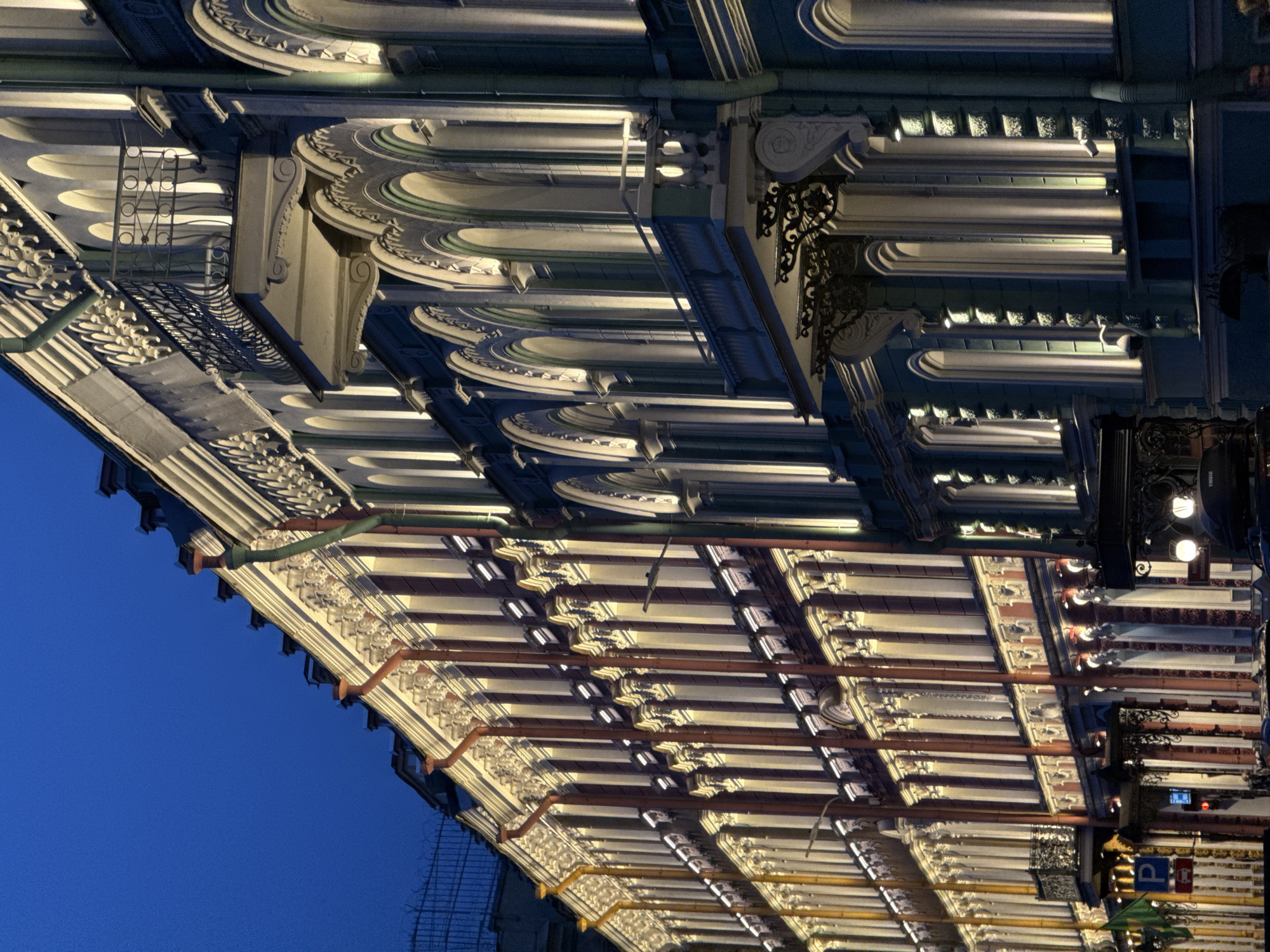 The image shows a low-angle, upside-down view of the ornate ceiling and supporting structure of a covered walkway or building, possibly a train station or a market. The architecture is characterized by intricate decorative elements, including sculpted cornices, arched recesses, and detailed metalwork. Linear lighting fixtures are integrated into the ceiling, casting a warm glow on the aged, likely stone or plaster surfaces. The overall impression is one of grandeur and historical significance.
The time of day appears to be evening or night, indicated by the artificial lighting and the dark blue of the sky visible at the bottom left of the frame, suggesting an outdoor element under a clear sky. The weather is not directly discernible but the lack of reflections on the sky suggests it might be clear or at least not raining.
Several objects are visible: metal pipes, possibly part of a drainage system, hang from the structure. Decorative wrought-iron brackets and lamps are also present, adding to the ornate aesthetic. In the distance, on the right side of the image, there are glimpses of signage, including a blue "P" for parking and a red sign with a white border, suggesting a public area. The intricate patterns and detailed craftsmanship point towards a location with a rich architectural heritage. The upside-down orientation creates a disorienting yet visually striking perspective, drawing attention to the complex details of the overhead construction. The scene is static, with no discernible activity or people. The location context of Saint Petersburg, Russia, aligns with the imperial and ornate architectural style often found in the city.