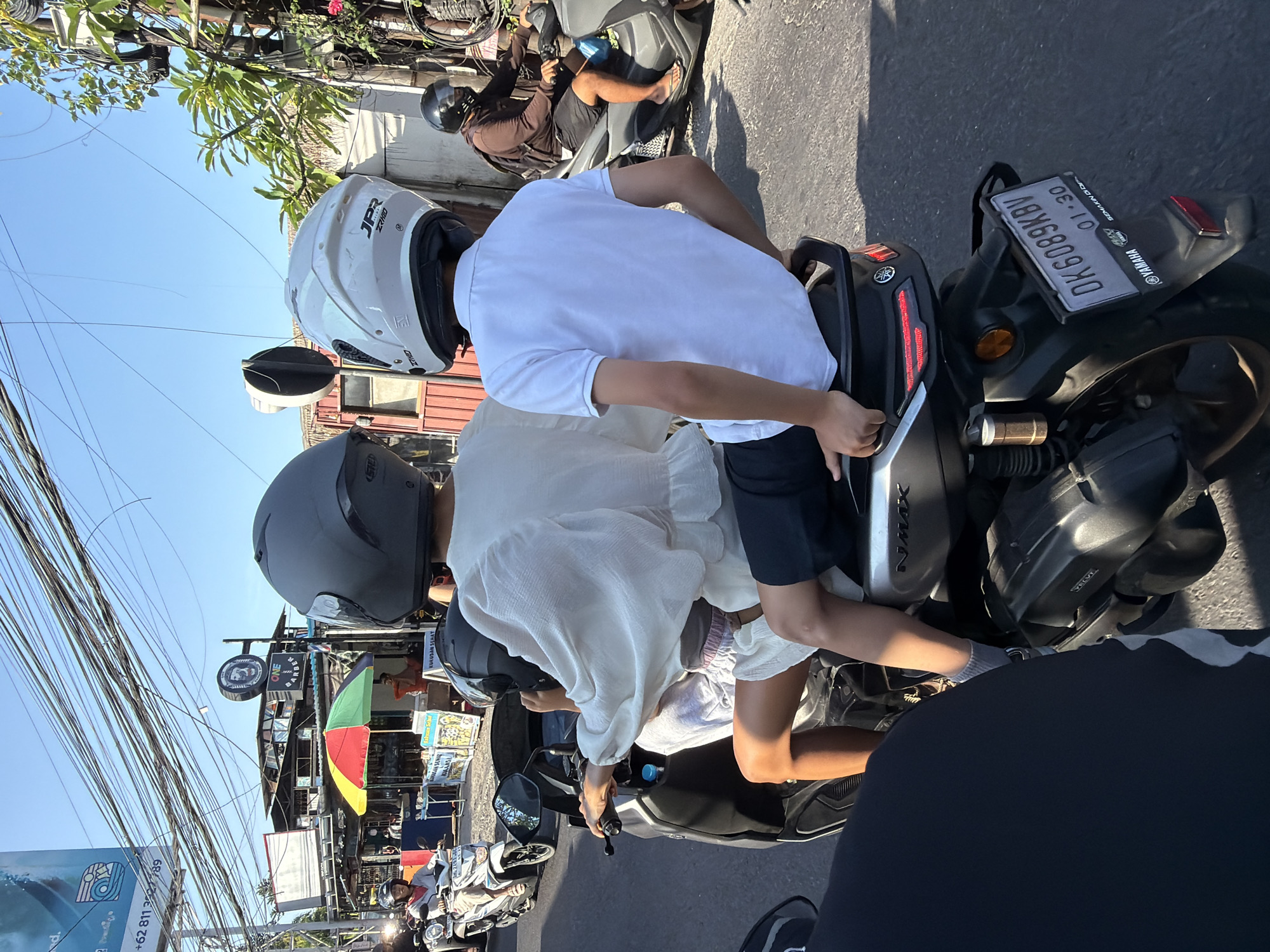 The image captures a street scene in North Kuta, Indonesia, under a clear blue sky. Two individuals are riding on a Yamaha Nmax scooter. The person in front, wearing a white polo shirt and black shorts, is driving, and their arms are visible gripping the handlebars. The person behind them, wearing a white puffed-sleeve top and white shorts, is seated as a passenger. Both individuals are wearing helmets. The scooter's license plate reads "DK 6089 KBV". In the background, there are various buildings, including a shop with a colorful umbrella and overhead wires, typical of an urban environment. Other motorcycles are also present on the road, and a person on another scooter is visible in the middle ground. The overall scene depicts everyday life on a busy street.
