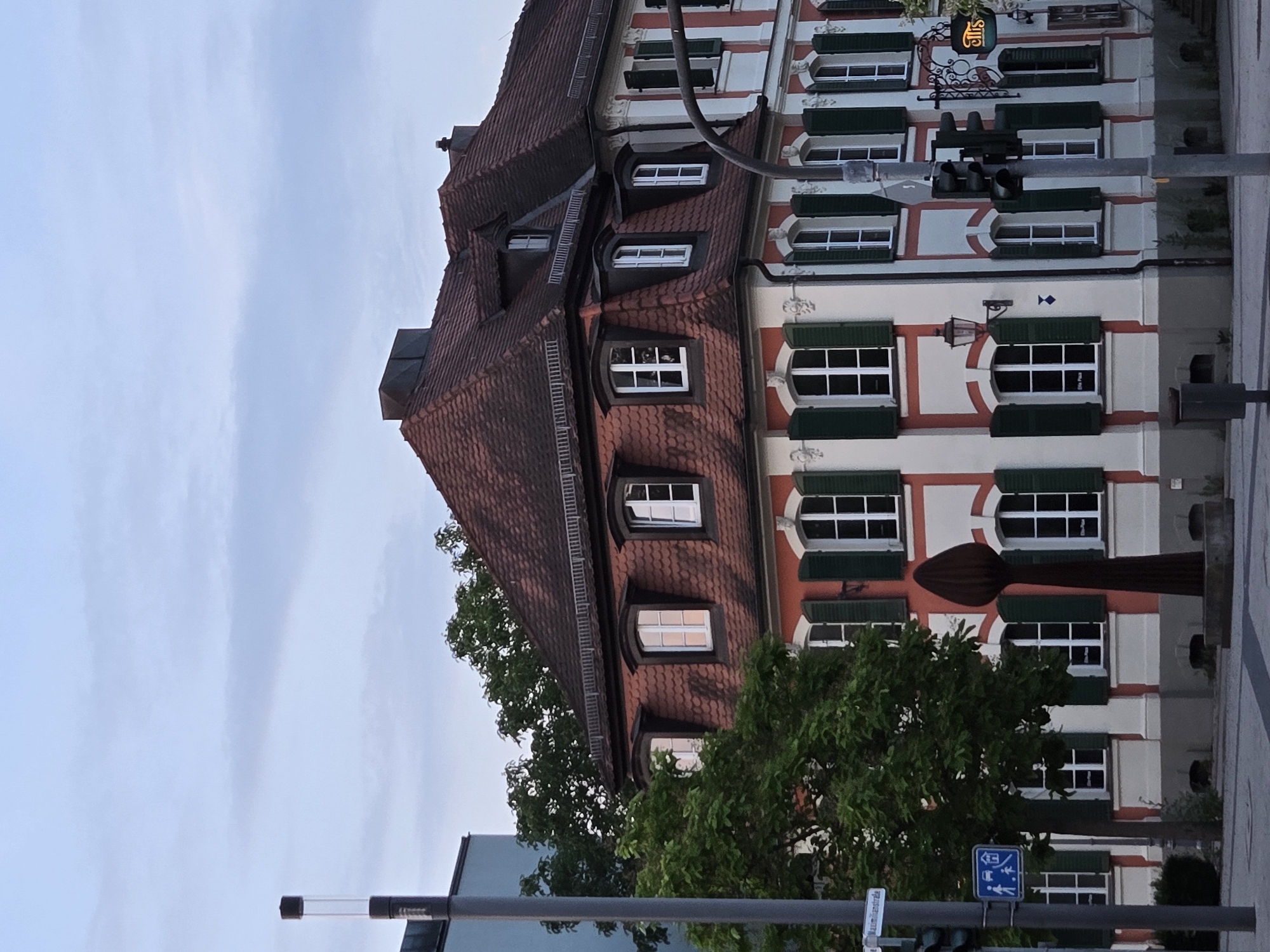 The image shows a corner building with a red-tiled mansard roof in Ansbach, Germany. The building has a white facade with red decorative bands and green shutters on the windows. There is a traffic light and a street sign visible on the left side of the image. A tree partially obscures the lower part of the building. A sculpture, resembling a stylized flame or seed, stands in front of the building. The sky is overcast with some clouds. The scene appears to be a street in a European city.
