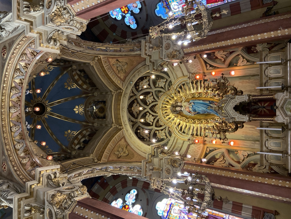 The image captures an ornate church interior from below, focusing on the ceiling. A statue of a woman with a radiating sunburst behind her stands out amidst intricate golden carvings and blue panels. Ornate chandeliers hang down, and stained glass windows add color. The architecture features arches and layered decorative elements, creating a visually rich and detailed space.