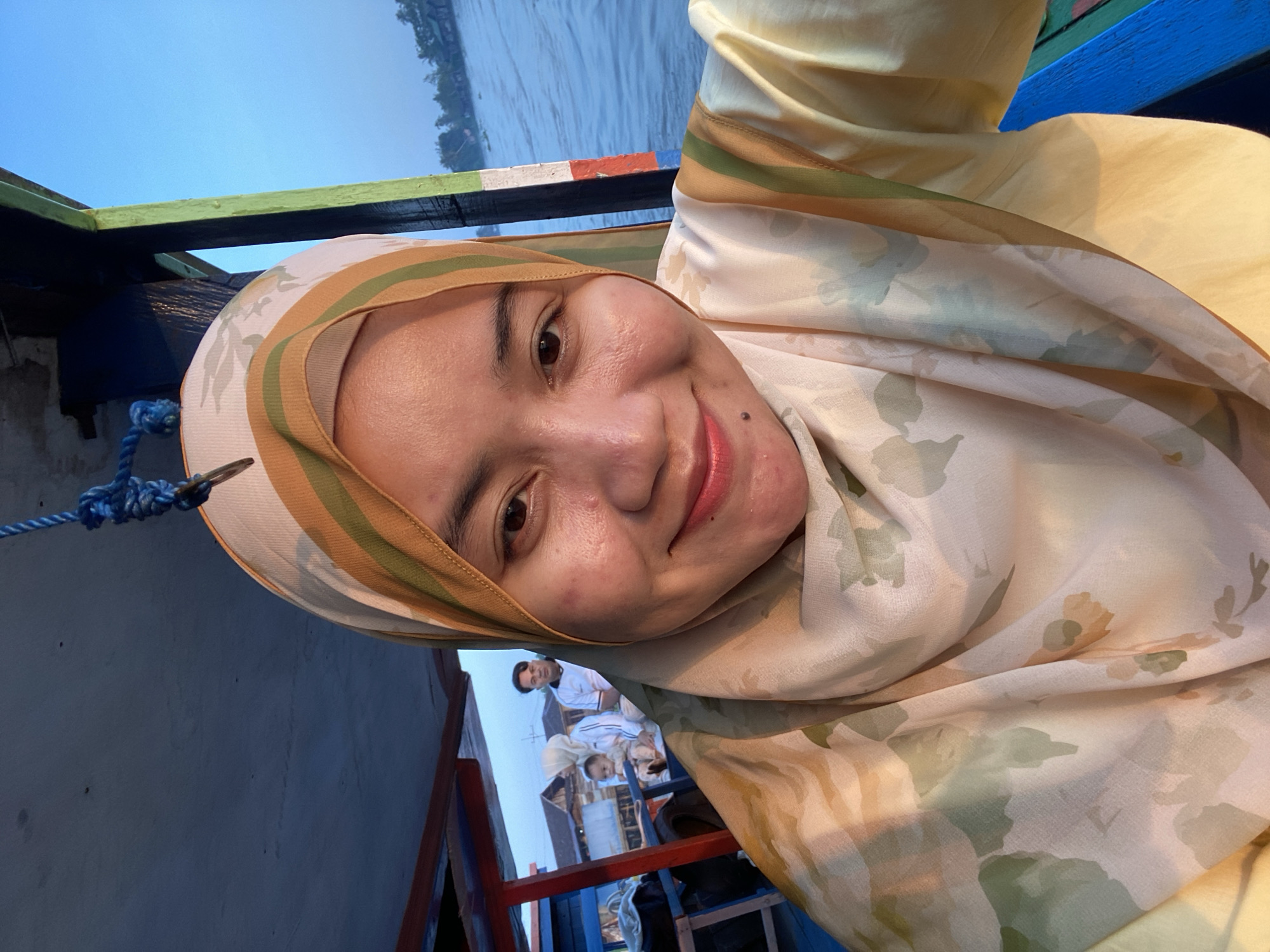A woman wearing a hijab and a floral patterned shirt smiles for a selfie on a boat. The boat's interior, with blue seating and wooden elements, is visible. In the background, other passengers are seated, and houses line the riverbank, suggesting the location is Sungai Tandipah, Indonesia. The water is calm, and the sky is a clear blue. The overall mood is relaxed and serene.