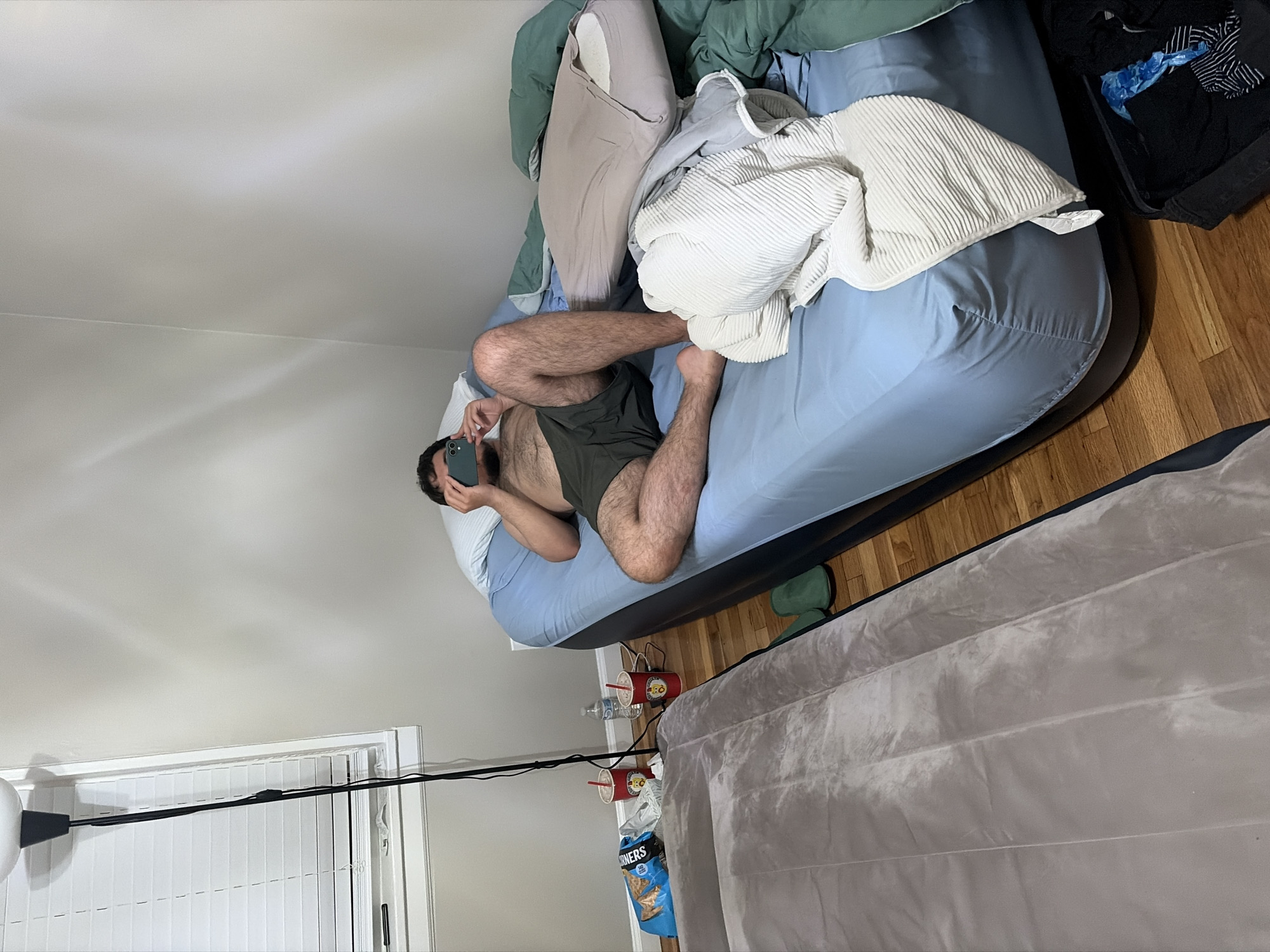A medium shot presents the interior of a room in Franklin Township, United States of America. Dominating the center is an adult male, reclining on a large, blue inflatable mattress. He is fair-skinned, dark-haired and bearded, and is wearing dark-colored shorts. He is angled towards the viewer, and is looking at a dark-colored smartphone in his hands. The mattress is covered with a pale blanket.
Around the mattress, a variety of objects are visible. A black bag filled with dark clothes sits in the corner. Beside the mattress, several red cups and a water bottle are visible. Next to this cluster is a gray air mattress. White window blinds are visible on the left side of the frame.