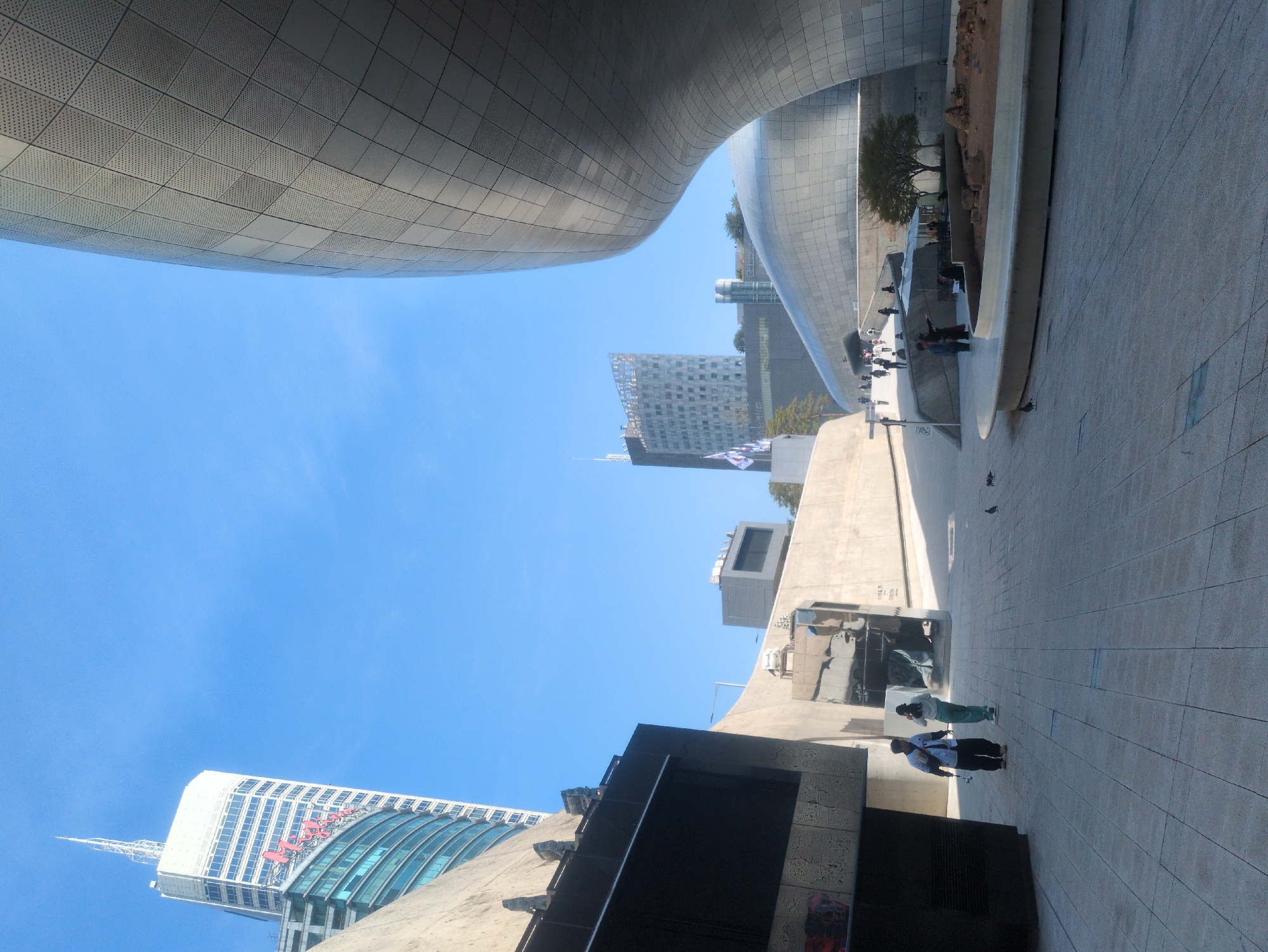 The image depicts an urban scene in Seoul, South Korea, featuring the Dongdaemun Design Plaza (DDP). The DDP's futuristic, curvaceous architecture dominates the view. The sky is a clear, bright blue. Several people are walking on the paved plaza. In the background, taller buildings are visible. The orientation of the shot is rotated ninety degrees.
