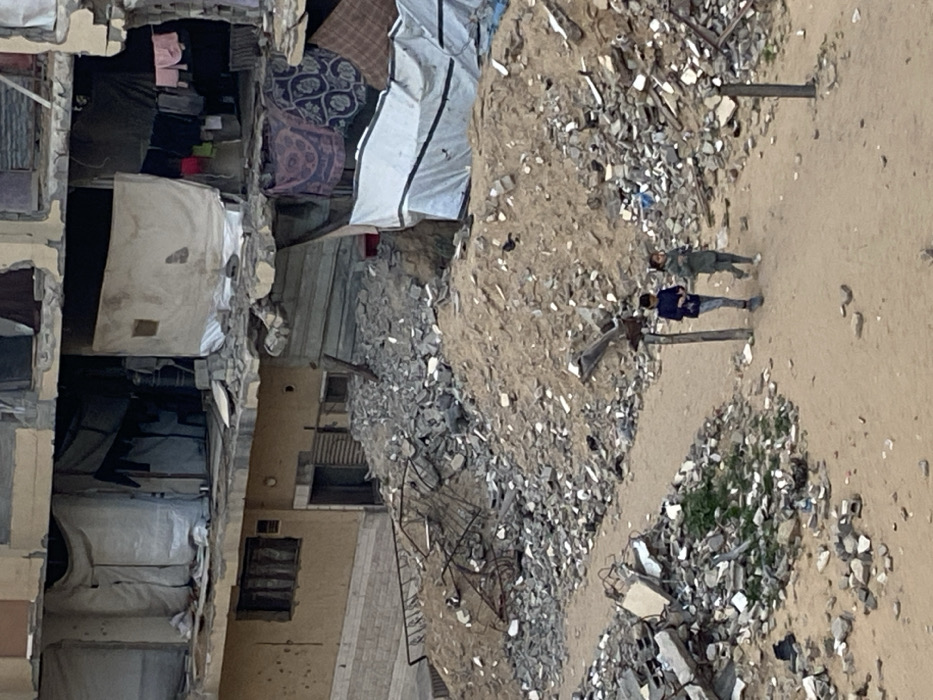 The image depicts a scene of significant damage and rubble, likely in an urban area, viewed from a slightly elevated perspective. The entire image appears to be rotated 90 degrees to the right, presenting a vertical orientation where the ground runs from top to bottom.
On the left side of the image, there are multi-story buildings, many of which appear heavily damaged or partially destroyed. The structures show exposed concrete, rebar, and broken walls. Inside some of the visible rooms, there are remnants of household items, such as what looks like bedding or blankets hanging, and possibly clothes. Tarpaulins or sheets are draped over some sections, possibly for temporary shelter or to cover damaged areas. One section of a building has a large, white, rectangular object that could be a door or a panel leaning against an opening.
The central and right portions of the image are dominated by uneven, dusty ground interspersed with large piles of rubble, consisting of broken concrete, bricks, and other debris. This suggests recent destruction or demolition. There are a few patches of green vegetation visible among the rubble, particularly in the lower right section.
In the middle-right of the image, two figures are standing on a relatively clear, sandy or dusty patch of ground. One person is wearing a dark blue top and light-colored pants, with their arms crossed. The other person is wearing green or camouflage-patterned clothing, possibly a military-style uniform. They appear to be observing the surroundings. Near them, there are a few wooden posts sticking out of the ground.
The lighting suggests it is daytime, with a soft, diffused light, possibly indicating an overcast sky or an early morning/late afternoon time. The overall impression is one of devastation and a possibly ongoing humanitarian situation, given the state of the buildings and the presence of people in such a setting. No discernible text is visible in the image.