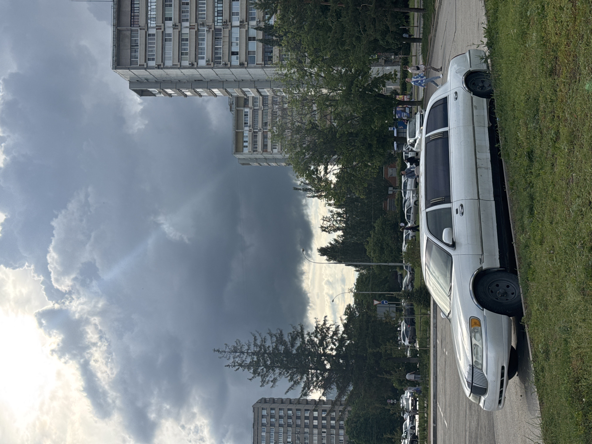 A white limousine is parked on a street next to a grassy verge. Other cars are parked behind it. In the background, there are apartment buildings and trees. The sky is filled with dark, ominous clouds. Two people are walking in the background. The location appears to be an urban area.