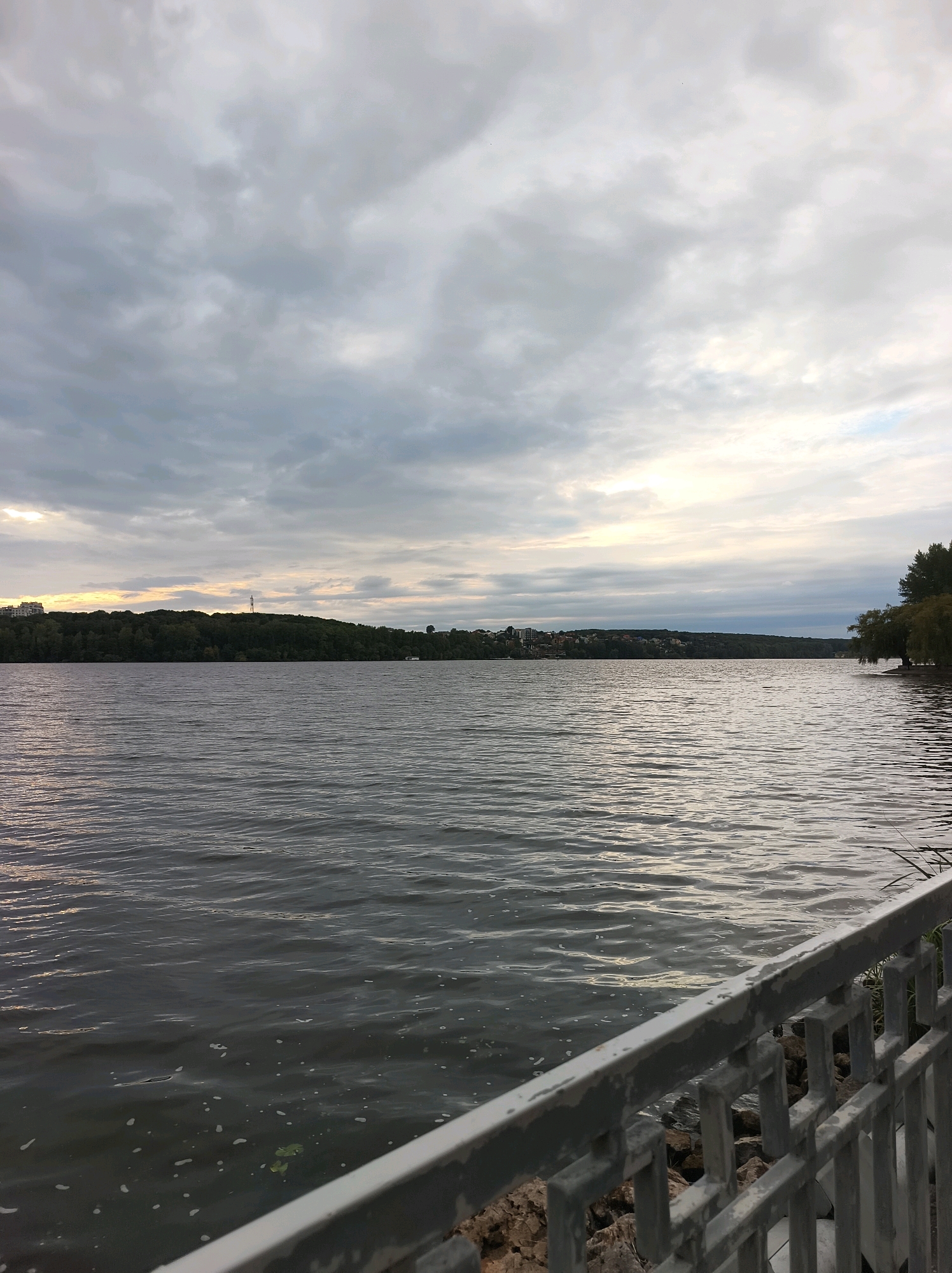 The image depicts a scenic view of Ternopil Lake in Ternopil, Ukraine. The foreground features a metal railing, partially obscuring a rocky shore. Beyond the railing, the calm surface of the lake stretches towards a tree-lined distant bank. The far shore is dotted with buildings and a distinct tower or monument on a hill. The sky above is overcast with dramatic clouds, hinting at approaching twilight or a stormy day, with a sliver of sunlight breaking through on the left horizon. The overall atmosphere is serene and natural, characteristic of a lakeside park or promenade.