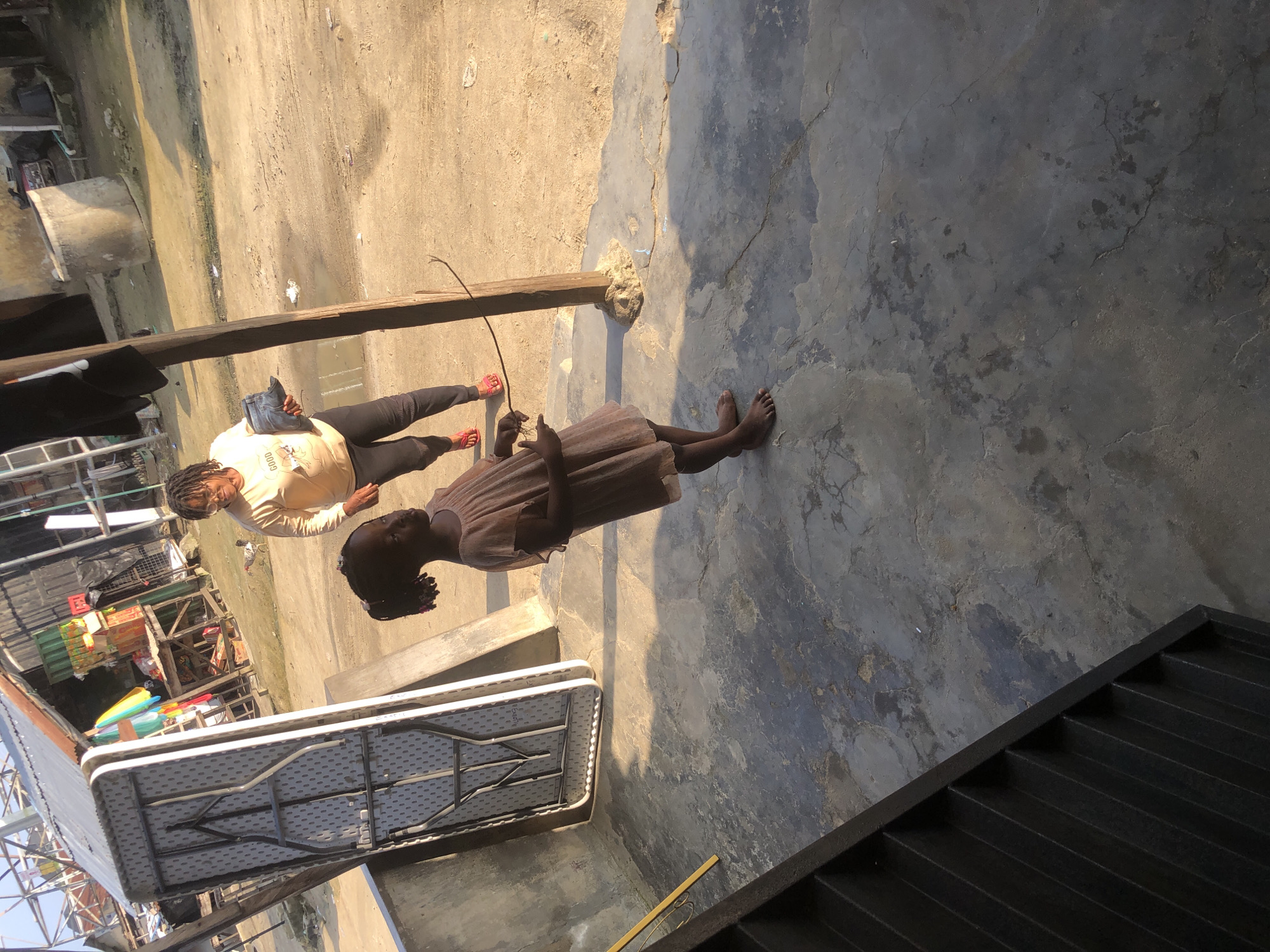 A young Black girl wearing a light brown, frilly dress stands barefoot on a cracked, concrete patio. She has dark hair pulled back into two pigtails and is looking to her right with her left arm extended. Behind her, a Black woman with glasses and braided hair wears a light beige t-shirt with a graphic on it and dark pants. She is holding a dark blue object in her right hand and walking towards the left side of the frame. A folded, grey table with a metallic frame sits on the patio to the left of the girl. The background shows a dirt road with a few buildings, some corrugated metal structures, and a large, cylindrical concrete water tank. A small chicken is visible in the middle ground of the dirt road. The lighting suggests it is late afternoon, with long shadows cast on the ground.
