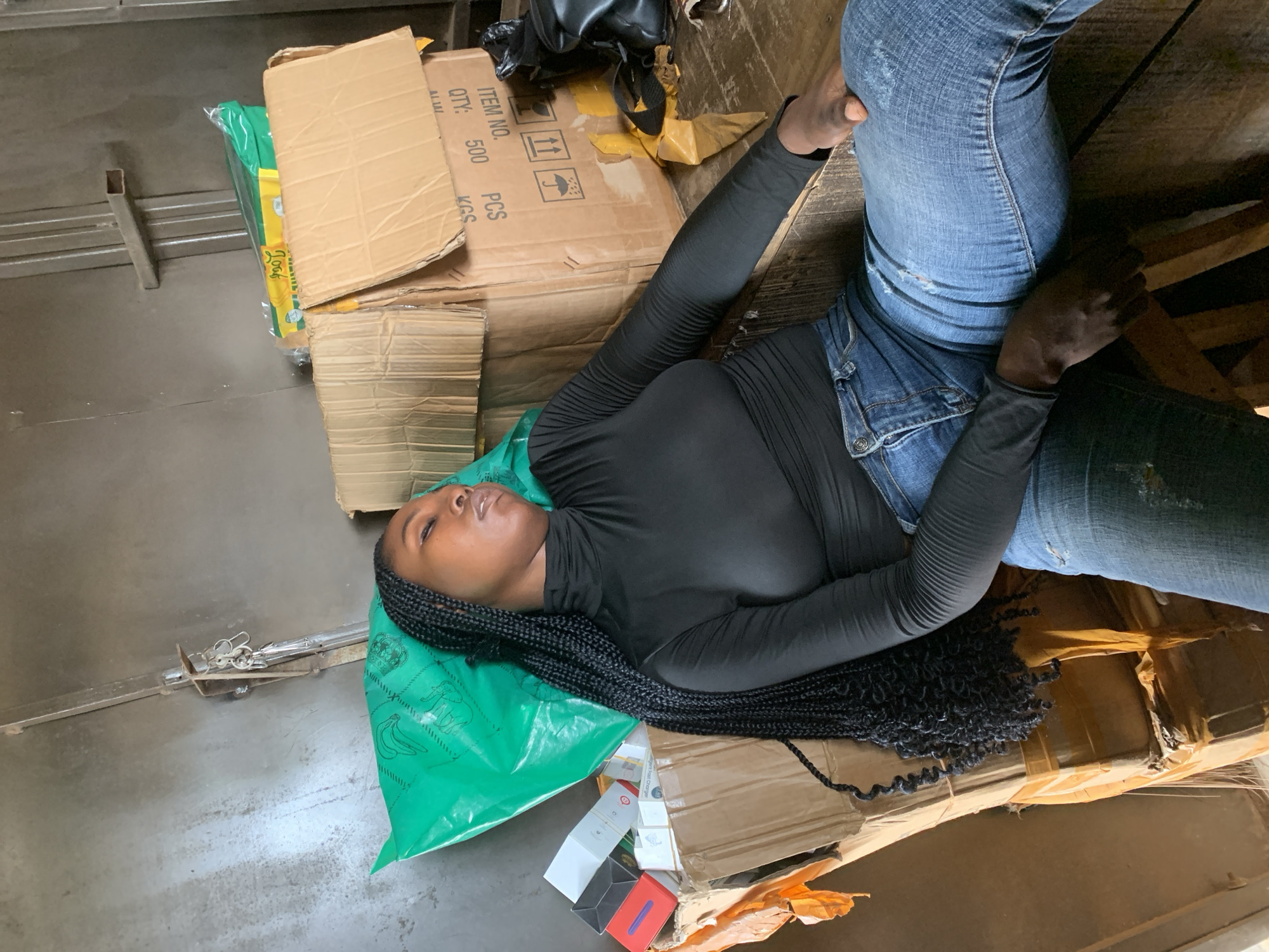 A young Black woman is lying down, asleep, on a stack of cardboard boxes in what appears to be a storage area or informal market stall. She is wearing a black turtleneck shirt and blue jeans, with her long braided hair spread out. The background consists of more stacked boxes and a metal structure. The scene suggests a place of commerce in Shagamu, Nigeria, where goods are stored and sold.