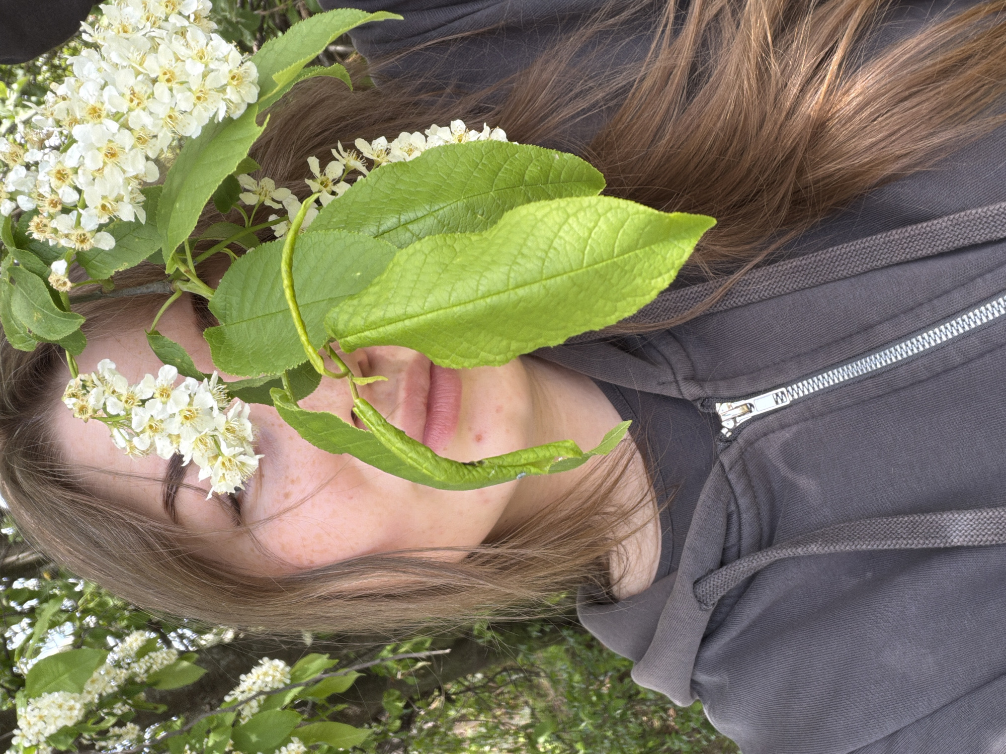 A young woman with brown hair is lying on the ground outdoors. Her eyes are closed. She is wearing a gray hooded sweatshirt that is partially zipped up. Her face is partially obscured by leaves and small white flowers. The background consists of green foliage and a glimpse of the sky. The location is Chaikovsky, Russia.