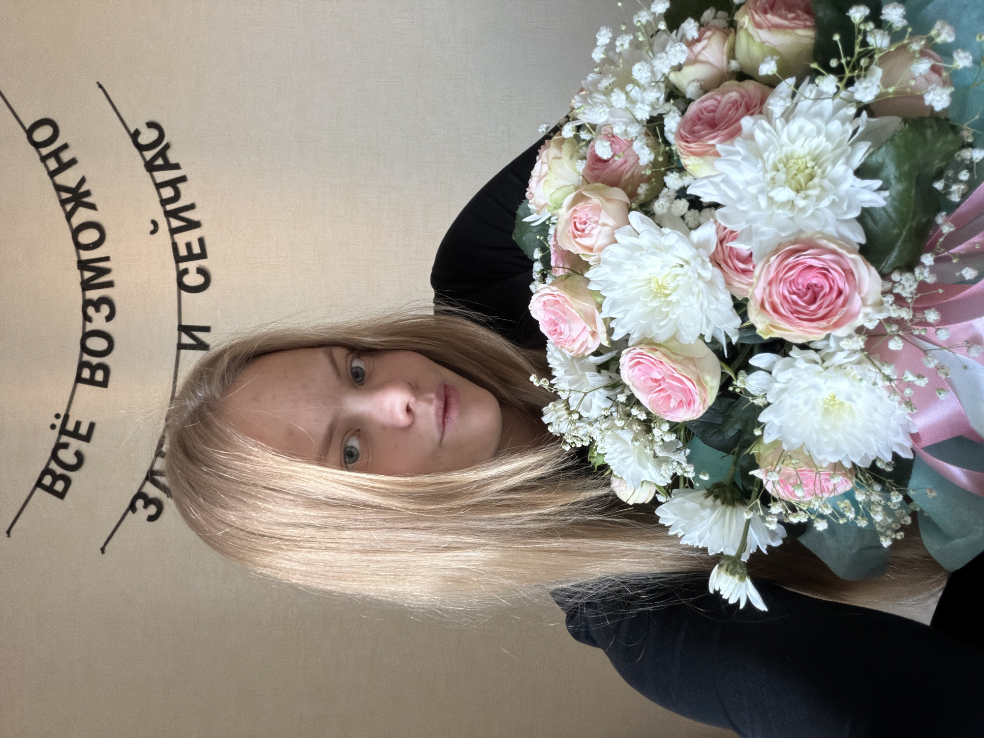 A young woman is captured in a close-up, slightly upward-angled shot, holding a vibrant bouquet of flowers. Her fair, shoulder-length blonde hair frames her face, and her light-colored eyes gaze directly at the camera with a neutral to thoughtful expression. She appears to be wearing dark or black clothing.
The prominent bouquet, held in front of her, features a mix of light pink roses, delicate white chrysanthemums, and airy white baby's breath, arranged with some green foliage. The flowers are wrapped in what appears to be light blue or teal paper and tied with a pink ribbon.
In the background, a light-colored wall or canvas is visible, displaying text in Cyrillic. The partially visible text reads: "ВСЁ ВОЗМОЖНО ЗДЕСЬ И СЕЙЧАС" which translates to "EVERYTHING IS POSSIBLE HERE AND NOW." The setting appears to be indoors, likely in Izhevsk, Russia, given the provided location context. The lighting suggests it is daytime.