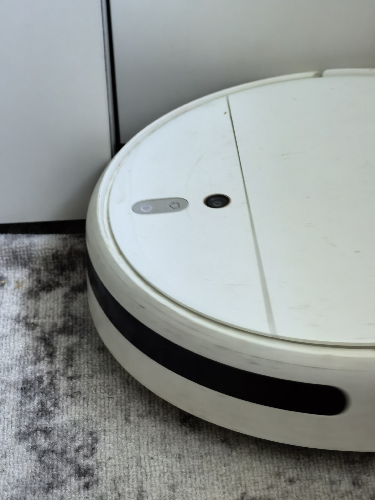 A white circular robot vacuum cleaner with a black stripe is positioned on a gray carpet next to a white cabinet. The robot vacuum has two buttons and a dark circular sensor on its top surface. The scene is indoors, with no indication of its specific location within the city of Sterlitamak, Russia.