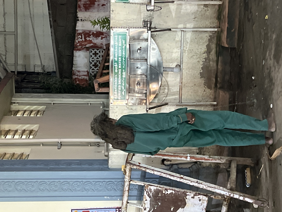The image depicts a person standing with their back to the viewer, next to a public purified drinking water dispenser, likely in Palani, India.
The central subject is a person wearing a plain teal-green two-piece garment, possibly a uniform or traditional attire, with long, dark, matted hair or dreadlocks extending down their back. A gold-colored ring is visible on a finger of their left hand, and their bare right foot is visible on the concrete ground.
To the right of the person, a stainless steel water dispenser unit is mounted against a wall. Above the dispenser, a green sign displays instructions in three languages. The prominent English text reads: "PURIFIED DRINKING WATER DON'T WASTE." Above the English, Tamil script conveys the same message: "சுத்திகரிக்கப்பட்ட குடிநீர் வீணாக்காதீர்" (Suththikarikkappatta kudiṉīr vīṇākkātīr), and below the English, Malayalam script also states, "ശുദ്ധീകരിച്ച കുടിവെള്ളം പാഴാക്കരുത്" (Don't waste purified drinking water).
The background shows parts of a building with light-colored walls and multiple horizontal slatted windows. Further right, a wall exhibits peeling reddish paint and features a white geometric lattice design. A small amount of green foliage is visible near this wall. The ground is a stained concrete surface. In the lower right foreground, a rusty metal frame or structure is partially visible. The lighting suggests daytime, possibly in a shaded area. The scene appears to be a functional public utility point, characteristic of a temple complex or public space in India.